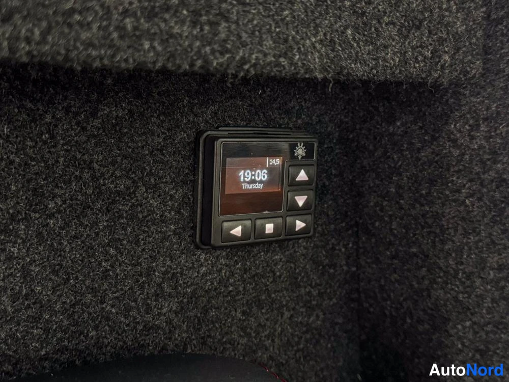 Yksityiskohta: A control panel mounted on the grey carpeted wall showing a digital display reading '19:06 Thursday' and a snowflake icon next to the number '14.5'. Buttons for