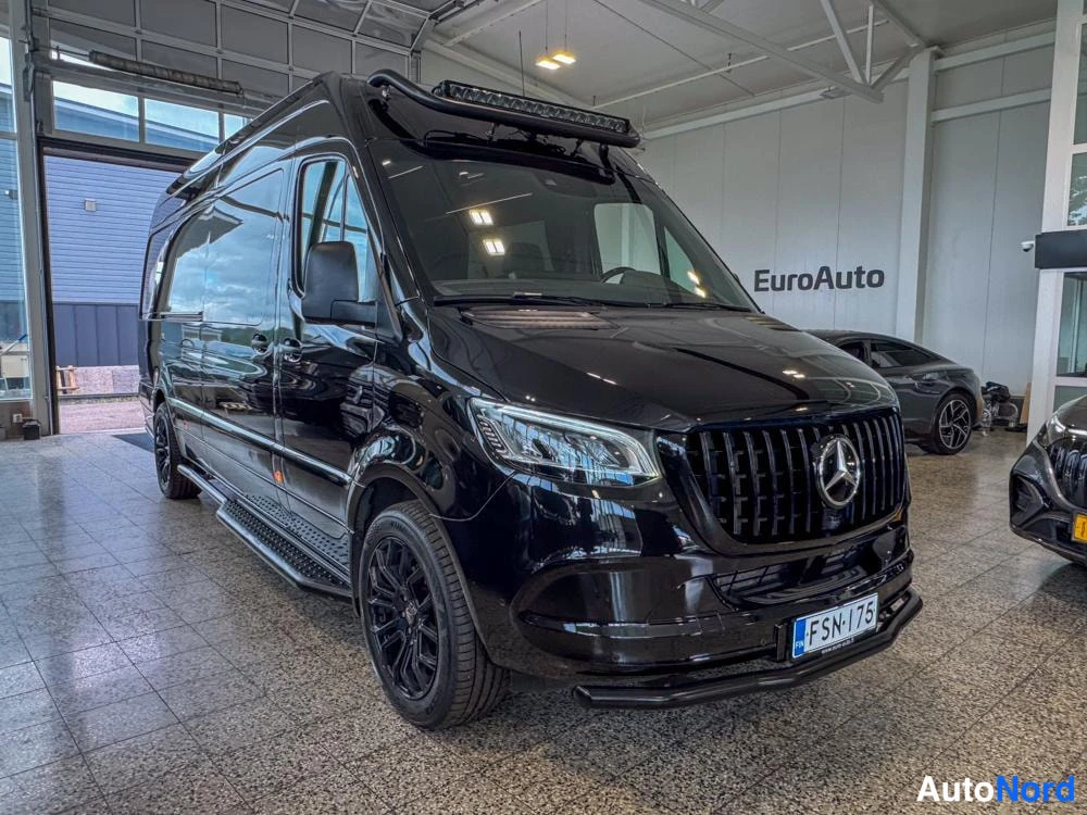 Etuviisto, nokka oikealle: Musta Mercedes-Benz Sprinter -pakettiauto etukulmasta autonäyttelyhallissa, nokka oikealla; katolla LED-valopalkki ja mustat vanteet.