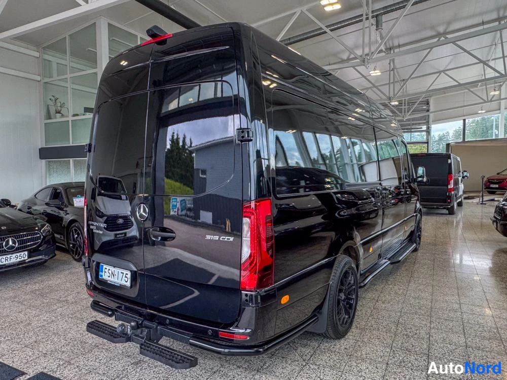 Takaviisto: Black Mercedes-Benz Sprinter van in a showroom seen from the rear right side, showing sliding door and towing bar.
