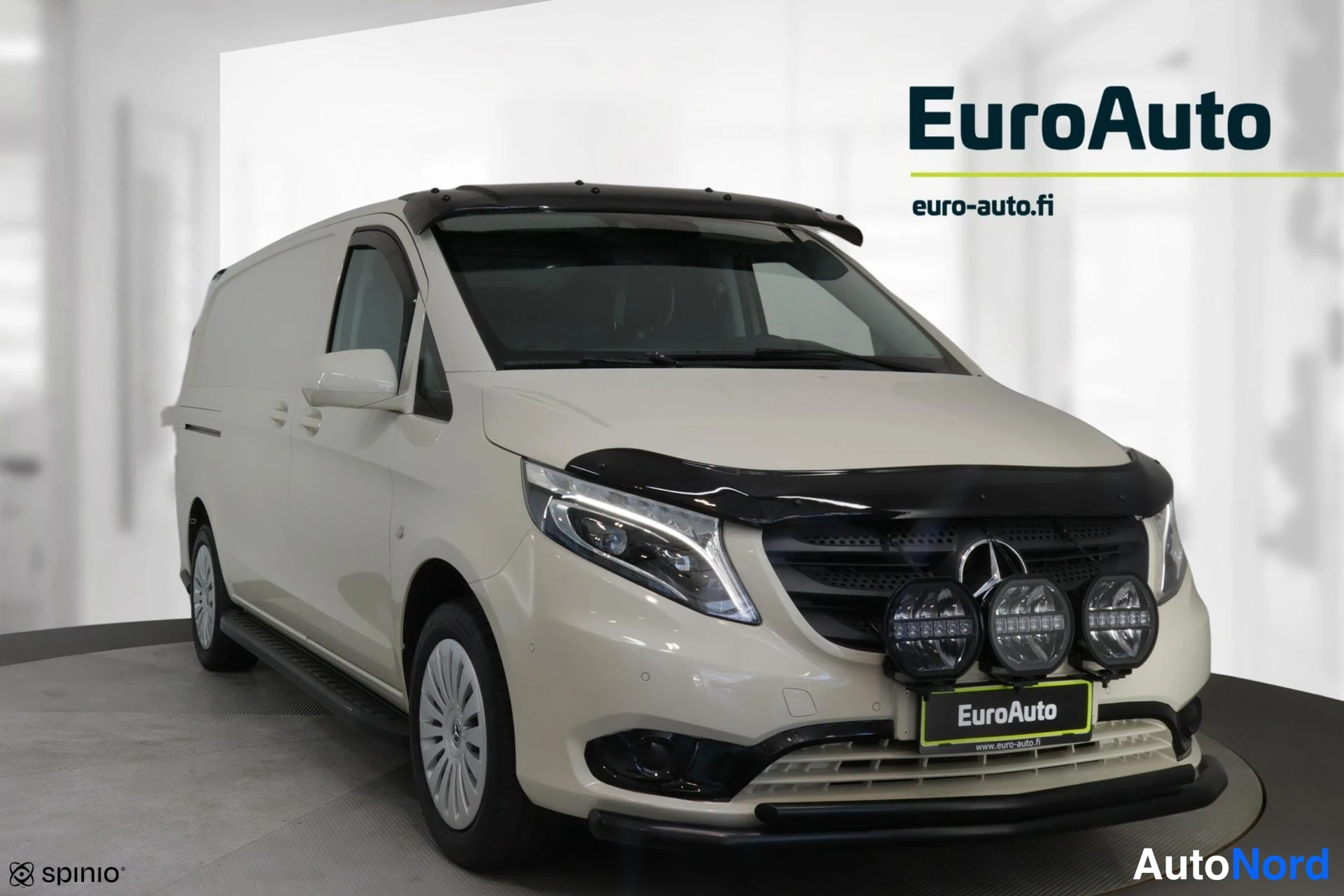 Etuviisto, nokka oikealle: Valkoinen Mercedes-pakettiauto kuvattuna etukulmasta oikealta; keulassa kolme pyöreää lisävaloa ja tumma suoja, katolla valopoimu, kyljessä lankut.