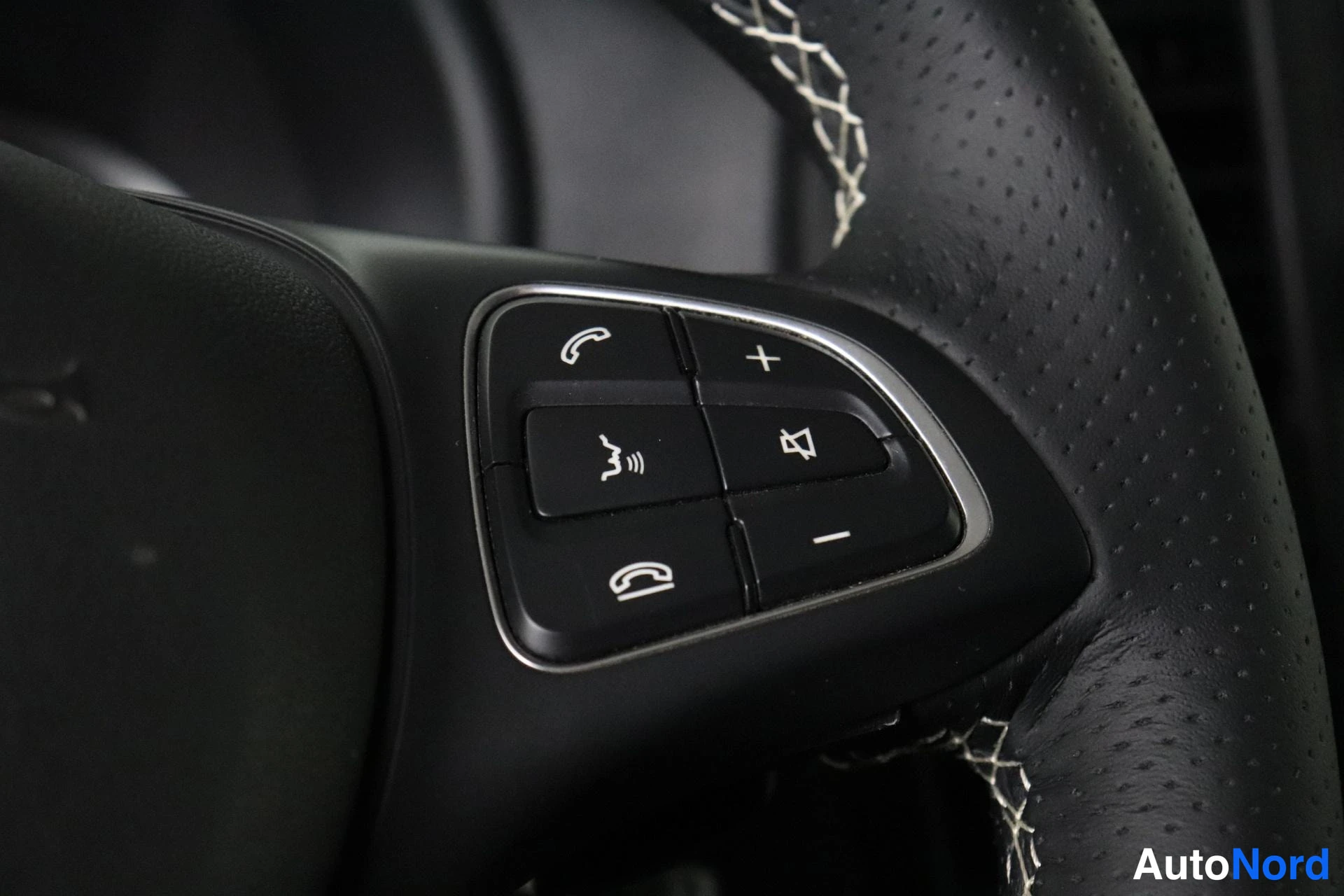Yksityiskohta: Close-up of the left side of a perforated leather steering wheel showing a control stalk with buttons for phone calls and volume adjustment.