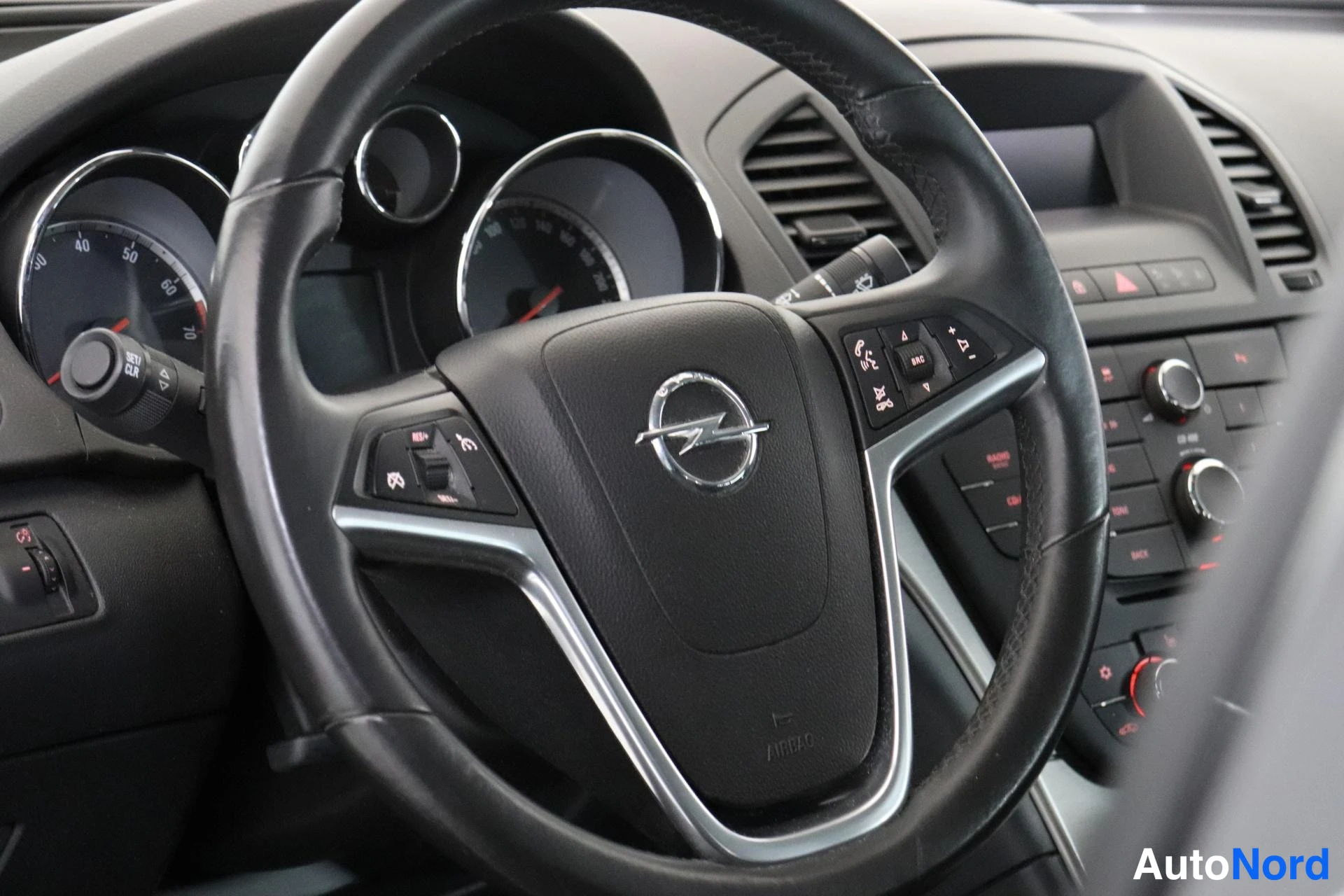 Etuviisto: Sisäkuva: Opel-merkkinen ohjauspyörä, jonka puoissa on monitoimipainikkeita; taustalla kojetaulun mittarit ja keskikonsolin radion säätimet.