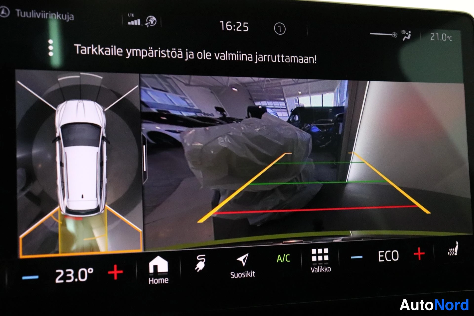 Yksityiskohta: Auton infoviihdenäyttö, jossa näkyy peruutuskameran kuva oranssi-, viher- ja punaisine ohjausviivoineen, ylhäältäpäin esitetty auton pysäköintitunnistinnäkymä s