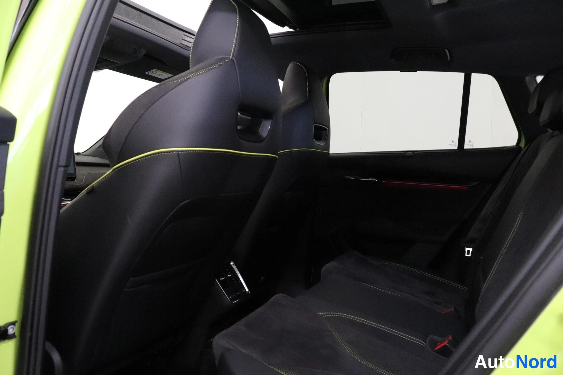 Takaistuimet: Rear passenger seats view from open rear door: black front seats with yellow contrast stitching; light green exterior door edge visible; ISOFIX child-seat ancho