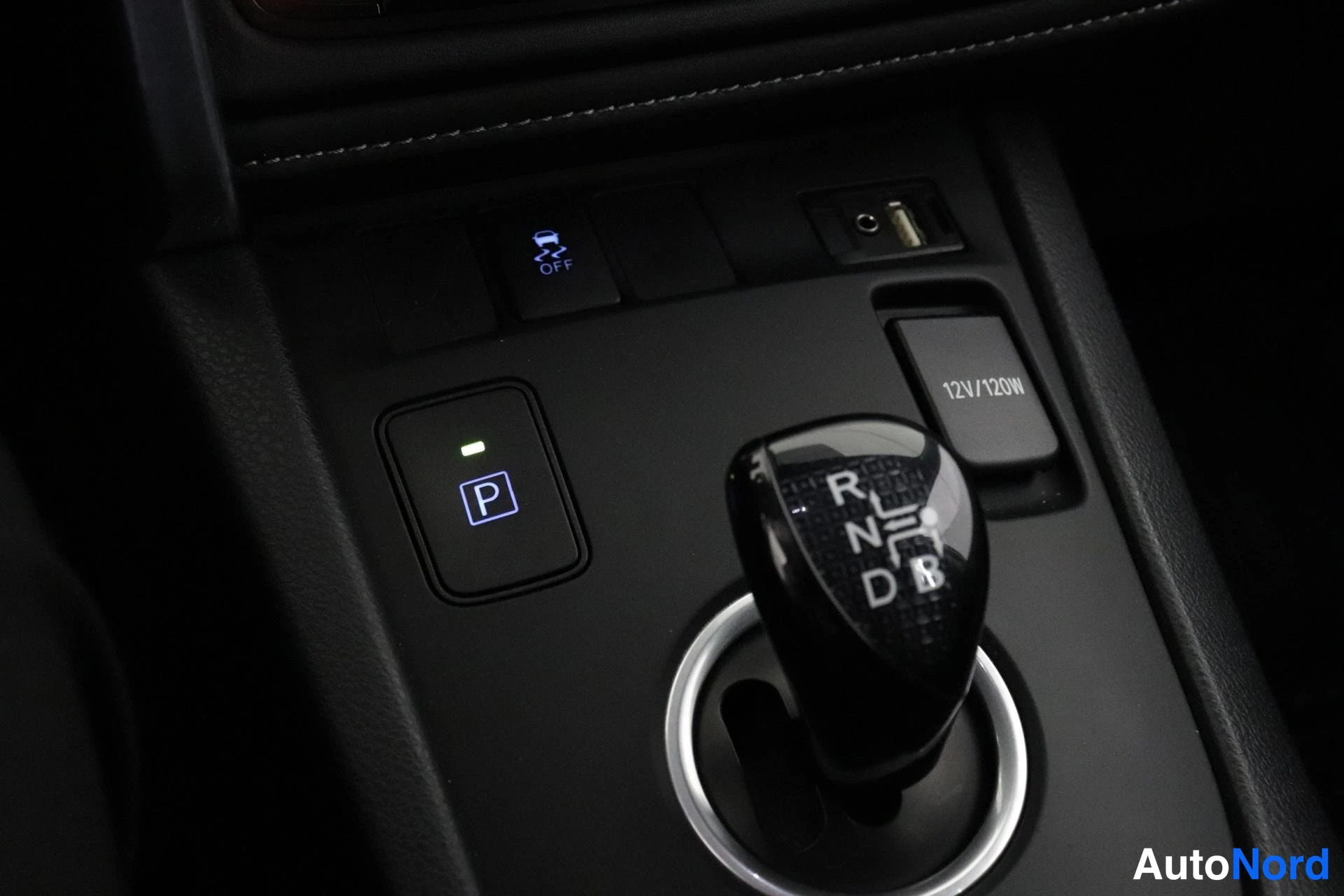 Yksityiskohta: Close-up of the automatic gear shifter in a car. A button with the letter P is visible next to the shifter.