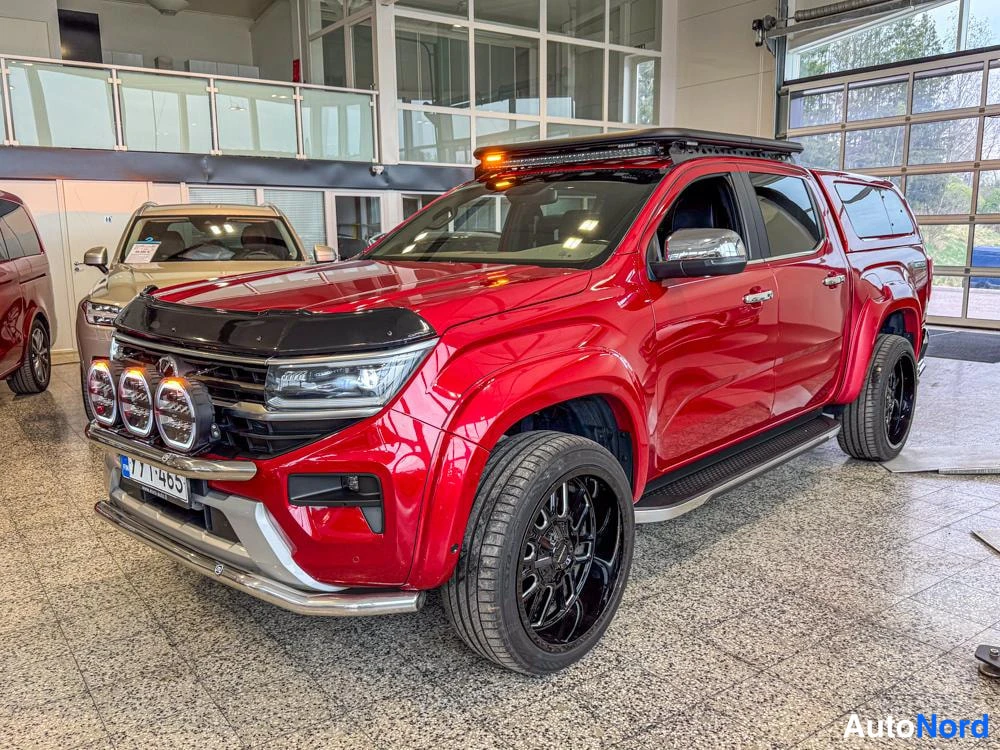 Takaviisto: Oikea edustakulma nähtävissä. Punainen Volkswagen Amarok, jossa katon päällä varoitusvalojen pitkä valo ja apuvalot etusäleikössä. Mustat vanteet ovat kiinni.