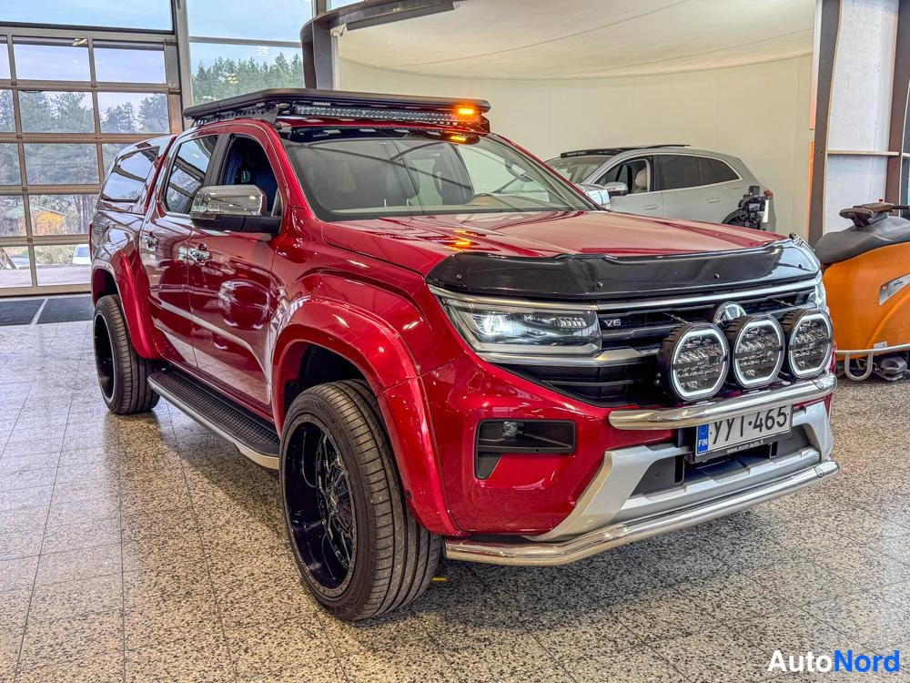 Etuviisto, nokka oikealle: Edessä oikealla kuvattu etuosa ja sivu. Punainen Volkswagen Amarok, jossa katon päällä apuvaloja, etusäleikön edessä apuvalot ja musta mytterin suojalevy