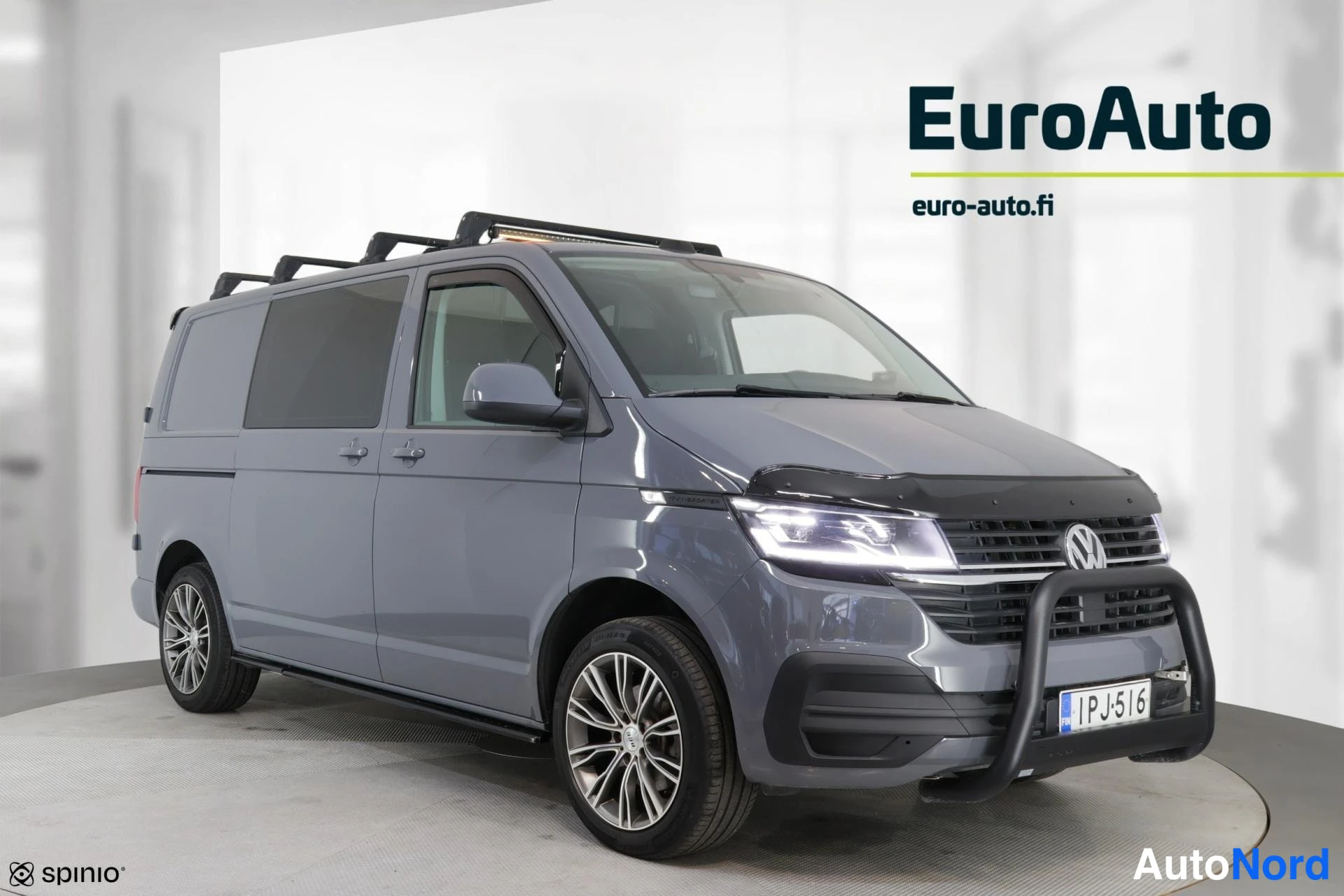 Etuviisto, nokka oikealle: Front three-quarter view of a grey van with LED daytime running lights on, roof rails visible, and alloy wheels. Grille is positioned to the right of the