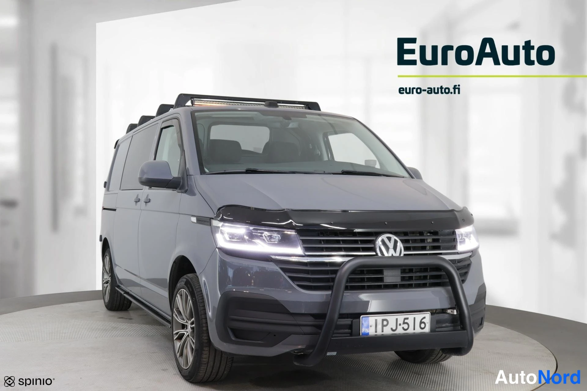 Edestä: Front view of a grey van featuring bright headlights, a chrome grille bar, a black bull bar, and a European license plate.