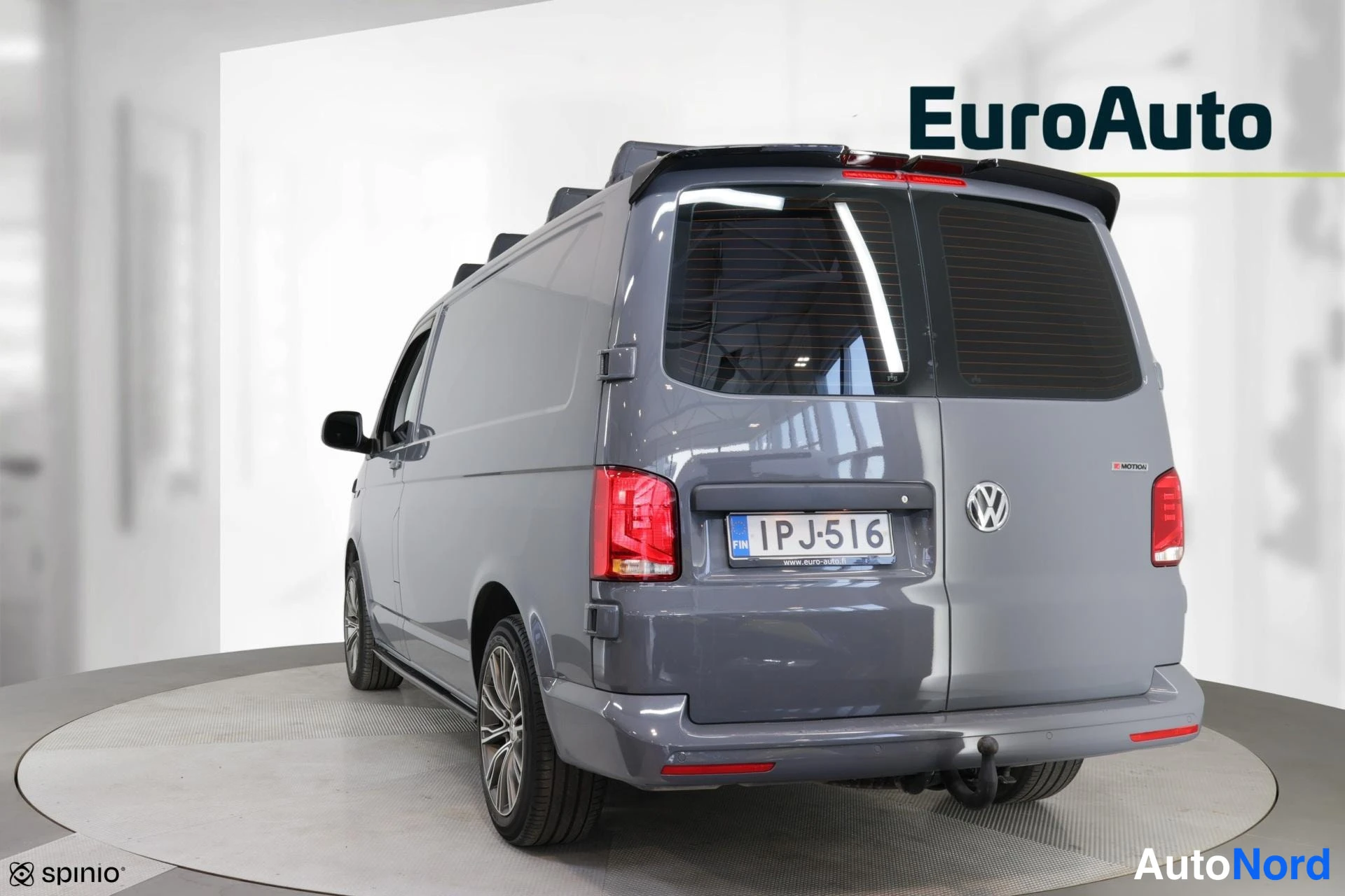 Takaviisto: Back and driver's side three-quarter view of a grey Volkswagen van with black roof rails, rear door featuring a VW emblem and license plate, visible tow bar, an