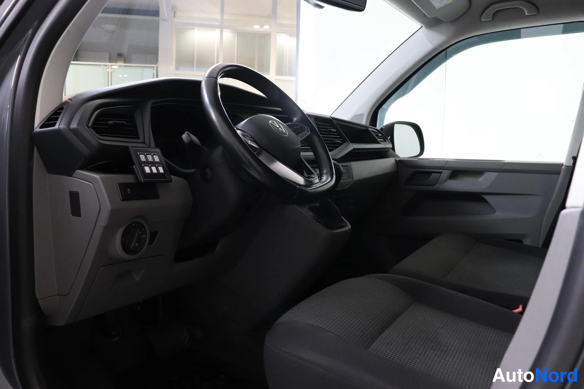 Ohjaamo: Interior shot from the open driver's door showing a black steering wheel with logos, dashboard, instrument cluster displaying '85', grey fabric front seats, man
