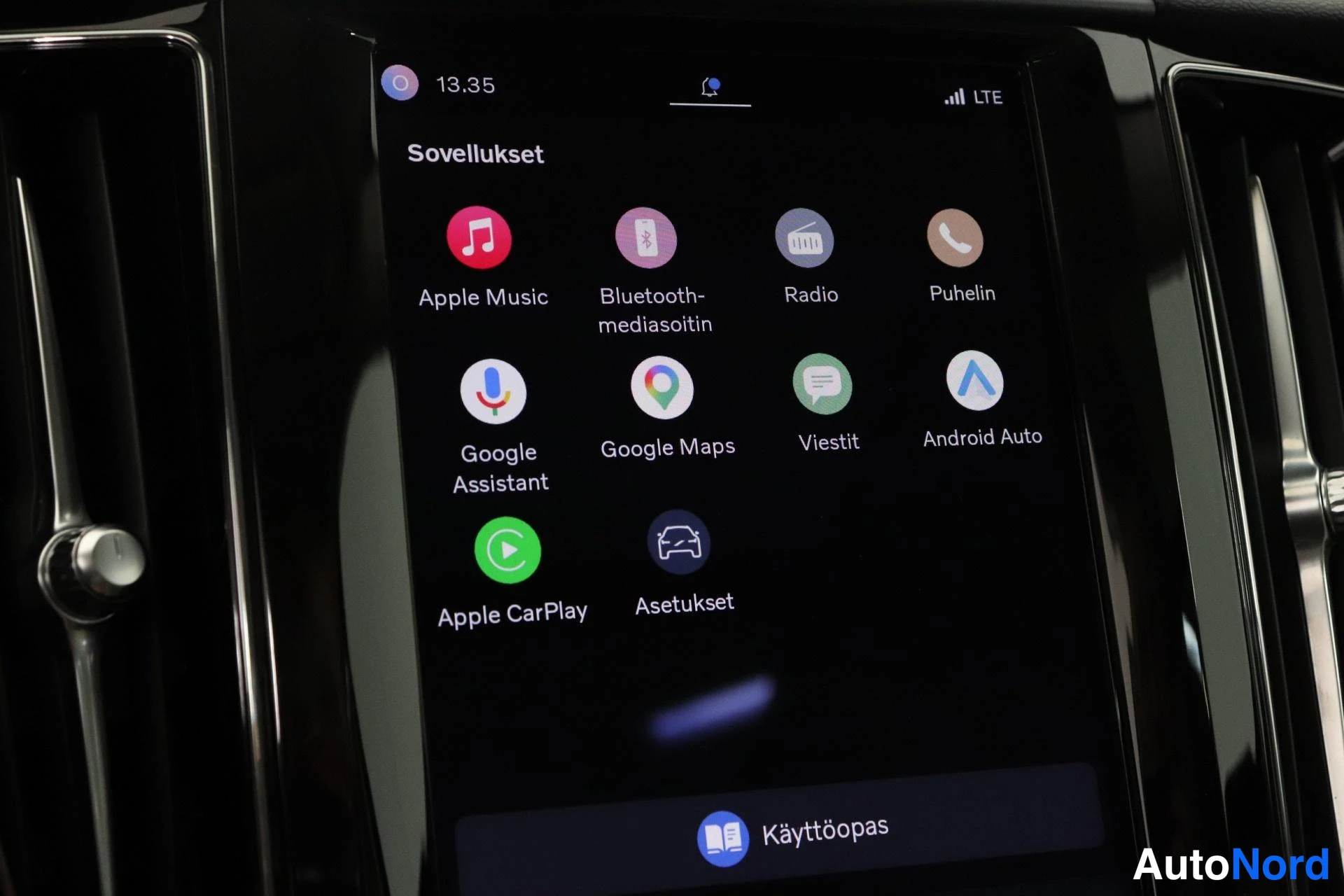 Yksityiskohta: Interior close-up of a large central infotainment touchscreen displaying the app menu. Icons for Apple Music, Bluetooth Media Player, Radio, Phone, Google Assis