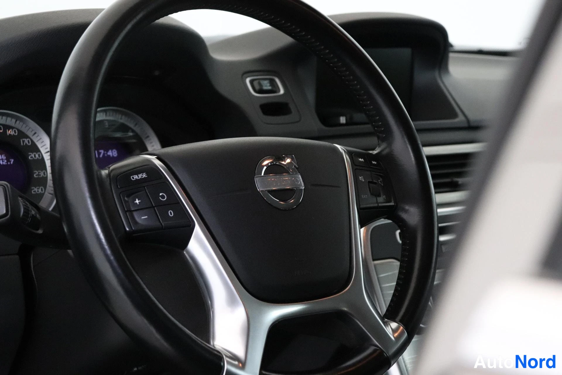 Yksityiskohta: Volvo-merkillä varustettu ohjauspyörä ja kojetaulu; vasemmalla CRUISE-painikkeet (+, −, 0 ja jatka-symboli), taustalla infotainmentnäyttö, nopeusmittari ja kell