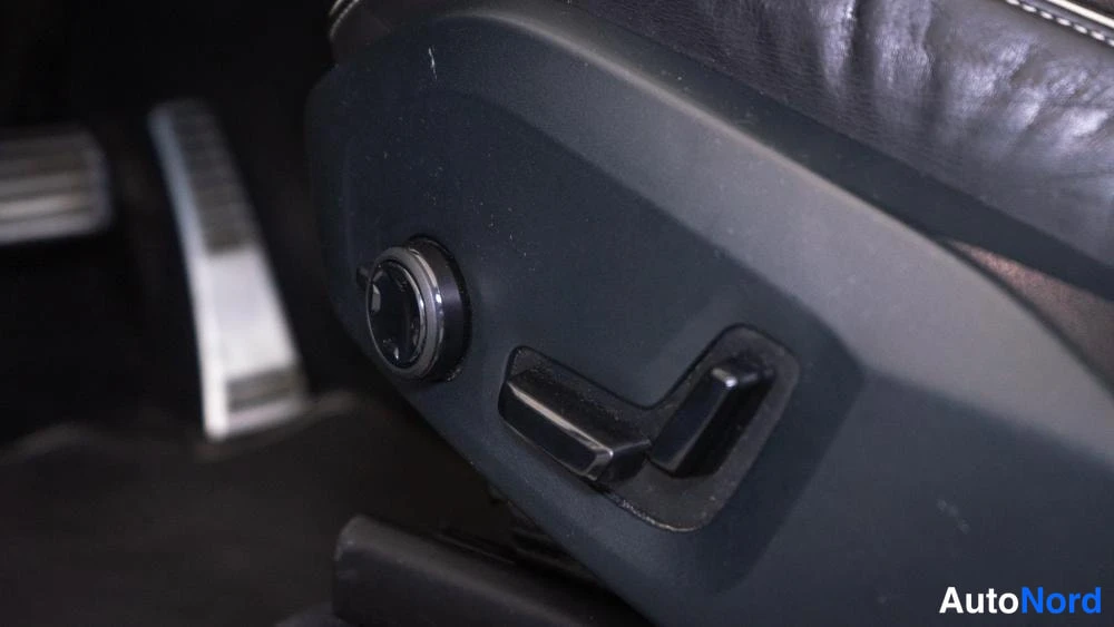 Yksityiskohta: Close-up of the side-mounted electric seat adjustment controls, featuring a circular rotary dial for backrest angle and an L-shaped toggle lever on the black pl