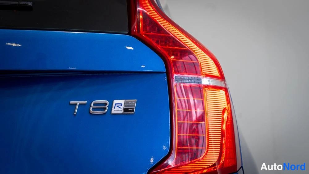 Takaviisto: Riittävästi taustavalossa oleva lähikuva auton takaosasta: sininen koristekyltti 'T8', pieni 'R DESIGN' -tunnus ja takaloistin oikealla puolella. Kuvan kulmassa