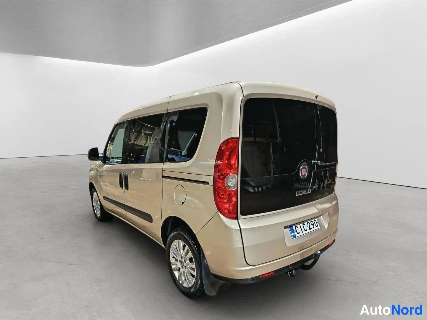 Takaviisto: Front left three-quarter view of a beige van displaying the driver's side profile, rear window, tail light, and alloy wheels, parked in a white studio.