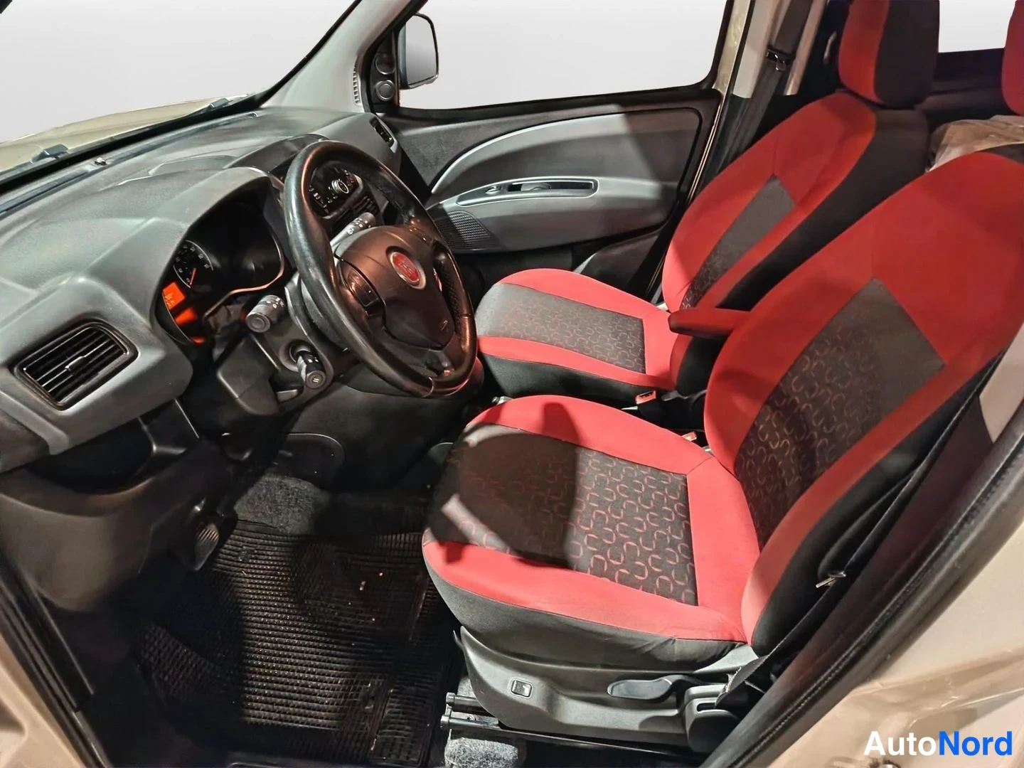 Sivulta: Auton sisätila: musta ohjauspyörä Fiat-tunnuksella, tumma kojelauta ja punamustat kuviolliset etuistuimet.