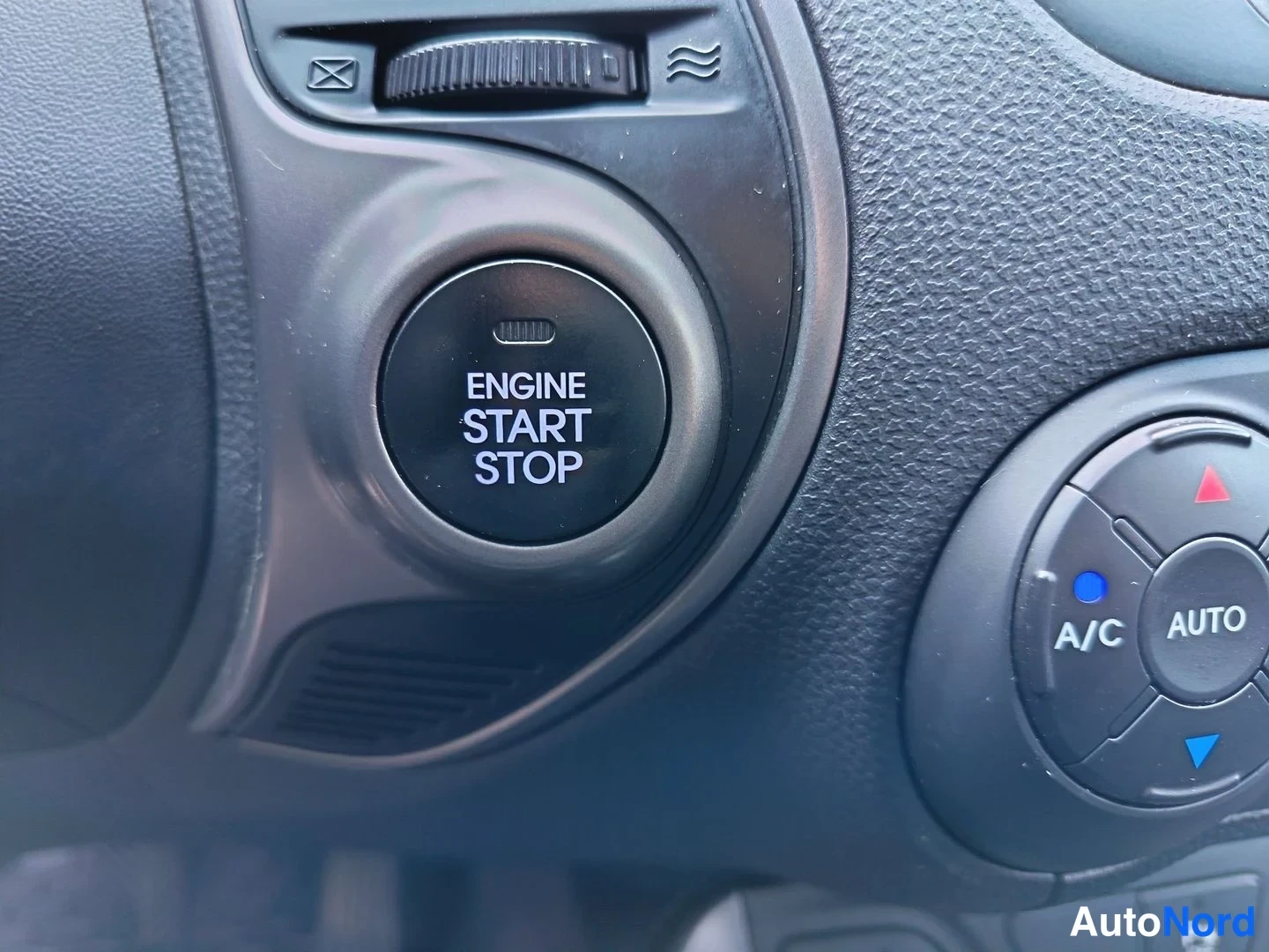 Yksityiskohta: Close-up of the vehicle's dashboard showing a circular ENGINE START STOP push-button ignition switch. To the right is the climate control dial with A/C and AUTO