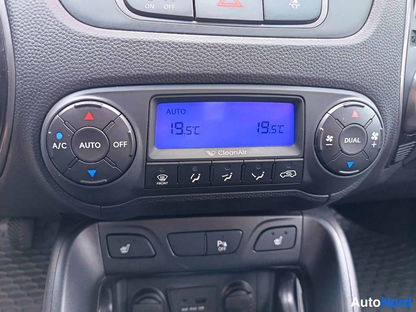 Yksityiskohta: Digital climate control panel with a blue backlit LCD displaying dual temperature settings of 19.5°C on both sides. Buttons for air distribution direction and d