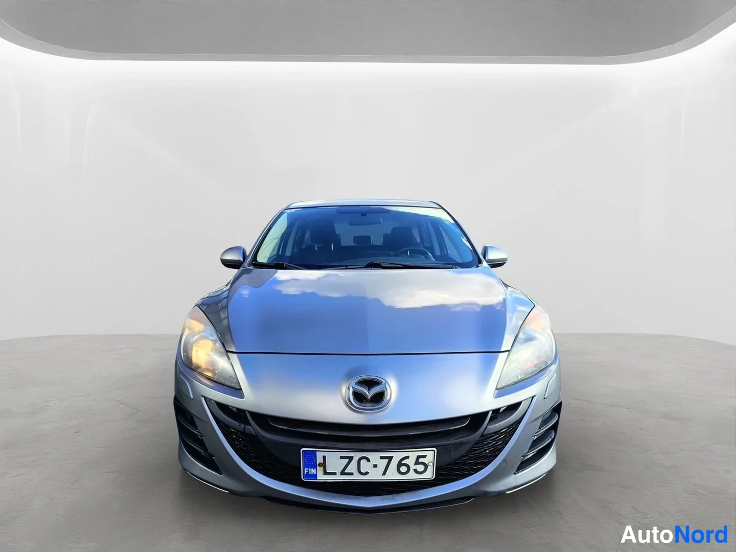 Edestä: Hopeanharmaan Mazda 3 -henkilöauton keula suoraan edestä, rekisterikilpi.