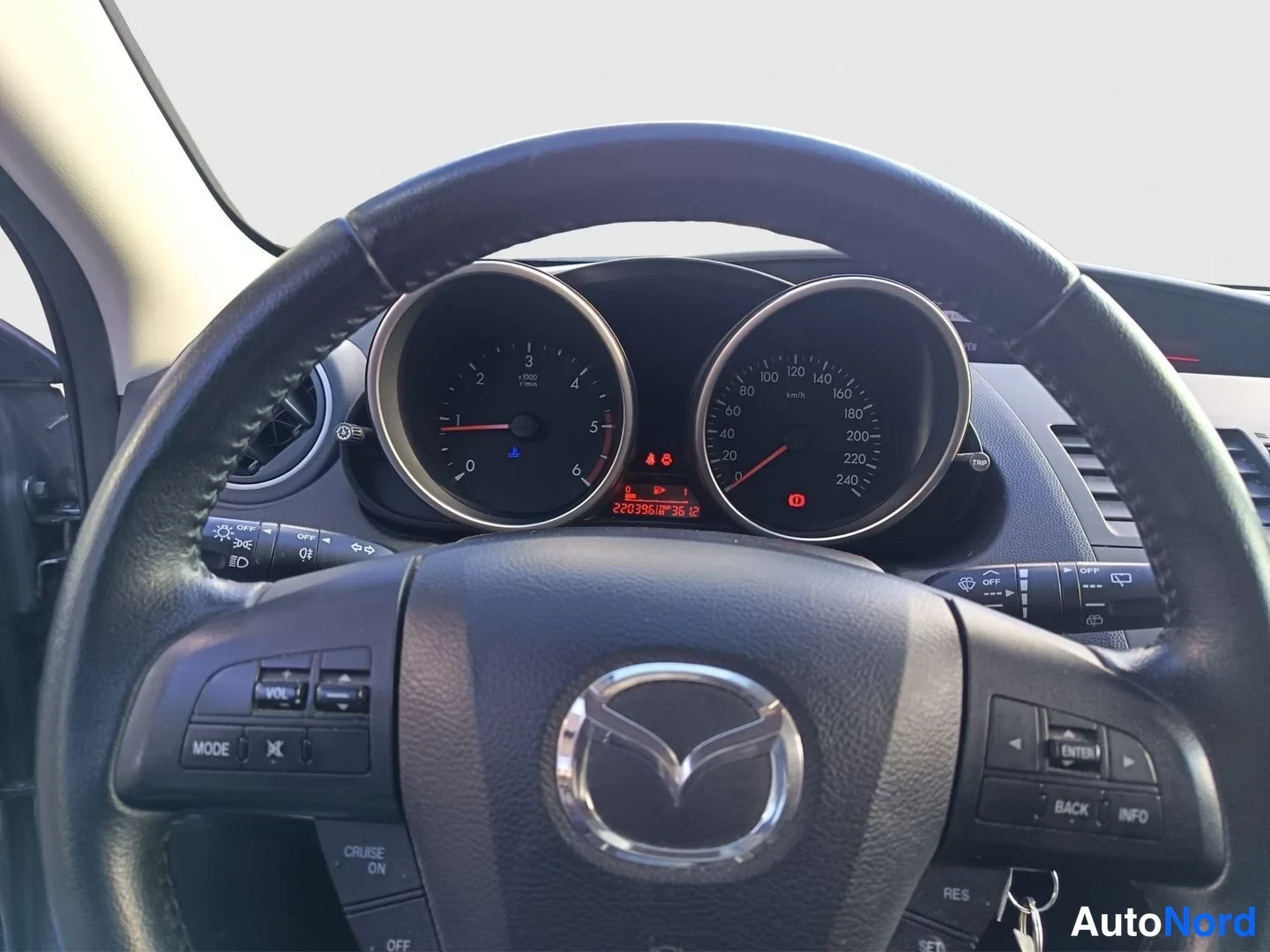 Ohjaamo: Close-up view of a black Mazda steering wheel and instrument cluster. The central digital odometer display reads 220398 km. A key is inserted into the ignition