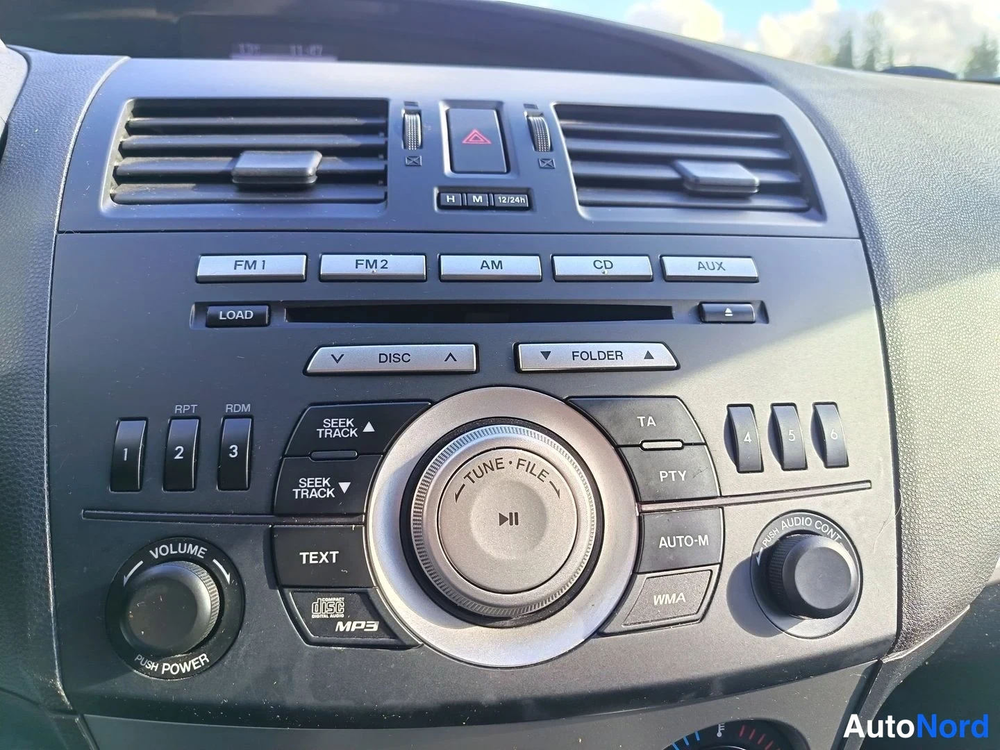 Yksityiskohta: Close-up of the car's dashboard audio unit. The panel features preset stations FM1/FM2, AM, CD, and AUX inputs. Controls include SEEK TRACK buttons, a large cen