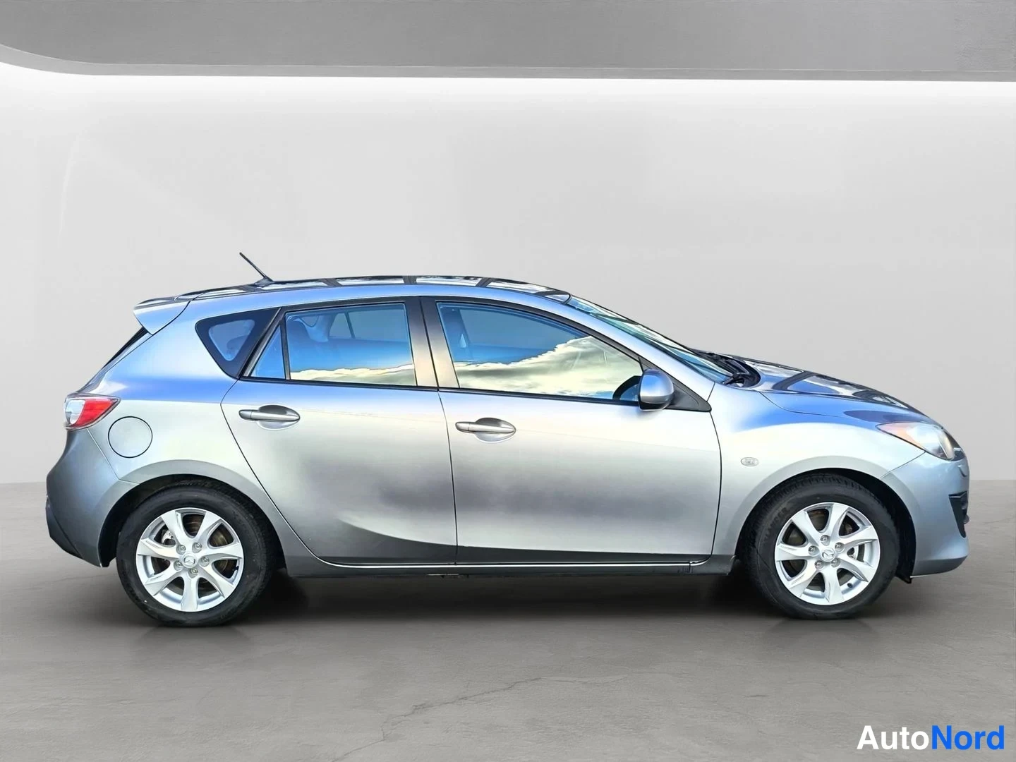 Sivulta: Hopeanharmaa Mazda 3 -henkilöauto kuvattuna suoraan sivulta, vasen kylki näkyvissä.