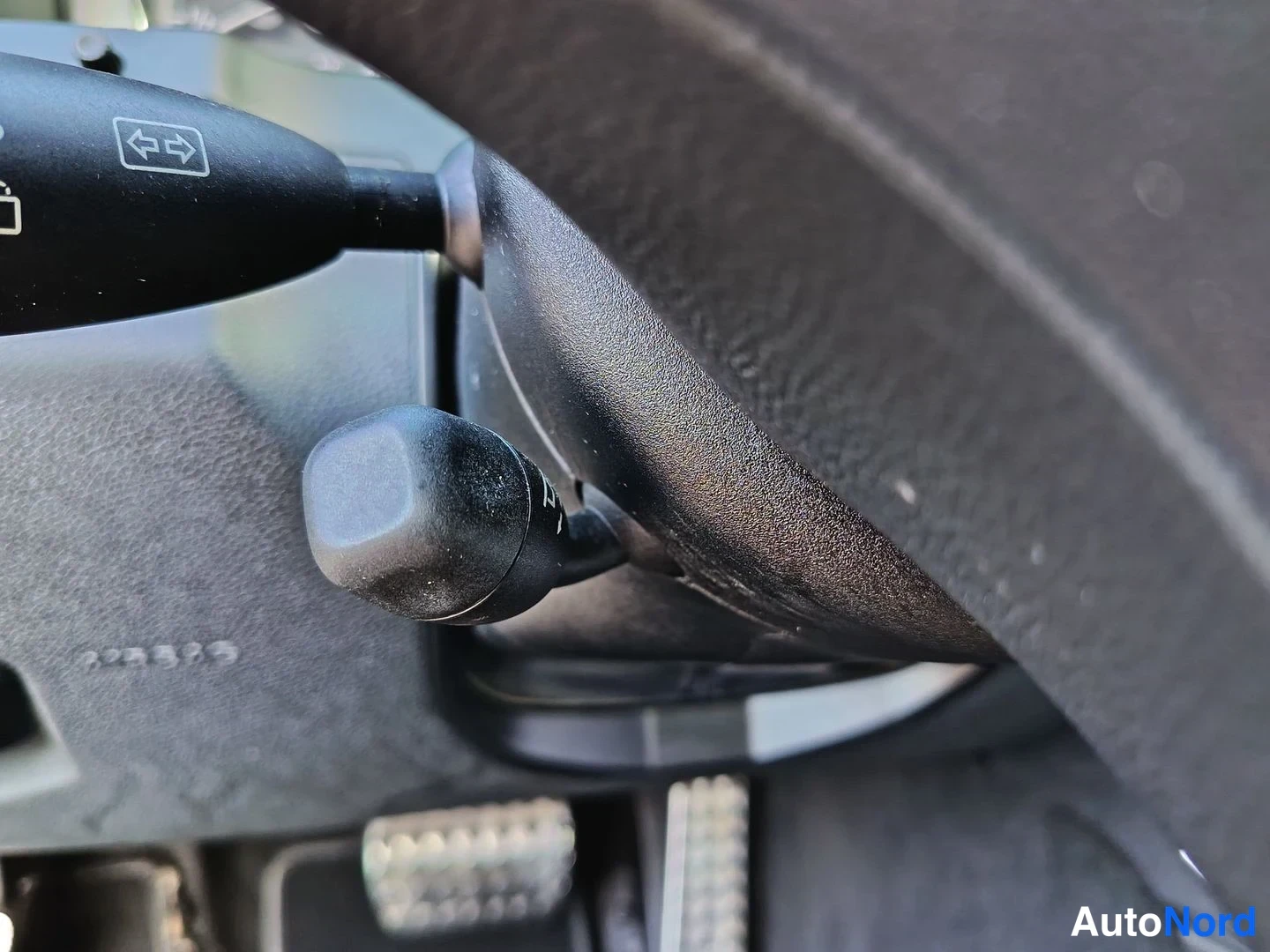 Yksityiskohta: Close-up of the right-hand stalk behind the steering wheel with a grey textured plastic knob; an indicator lever is visible to the left. A chrome foot pedal is