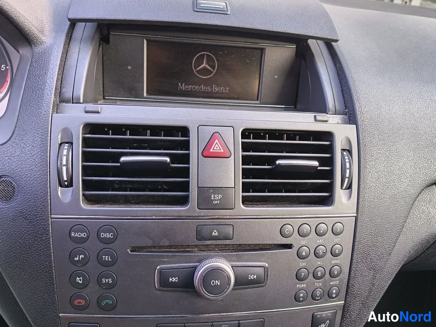 Yksityiskohta: Centre console showing a closed display unit with Mercedes-Benz logo above air vents and red hazard button; a radio control panel with 'RADIO', 'DISC', 'TEL' la