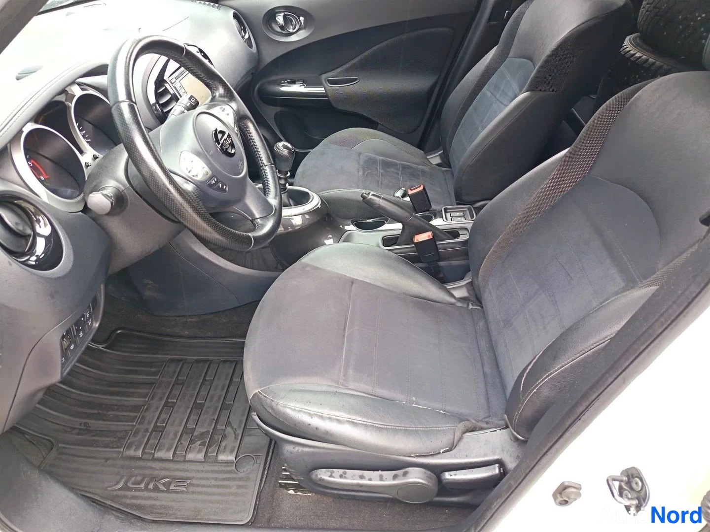 Muu: Nissan Juken sisätilat kuljettajan puolelta: manuaalivaihteisto, tummat penkit, JUKE-tekstillinen matto ja tumma kojelauta.