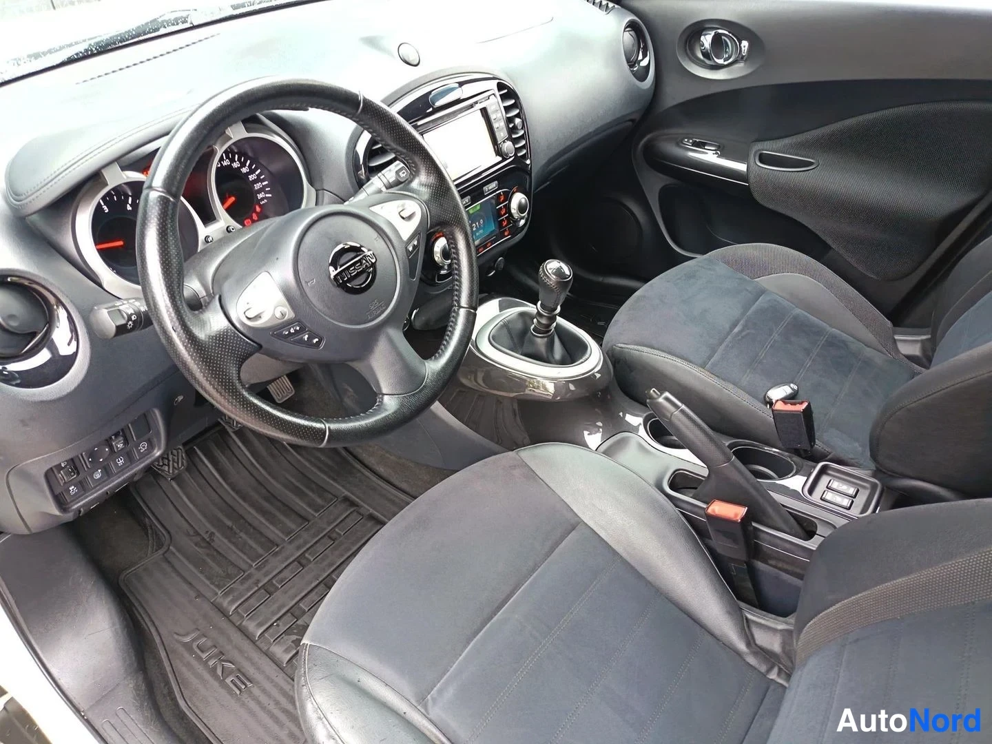 Muu: Ohjaamon näkymä: Nissan-ohjauspyörä, manuaalivaihtopyörä, tummat penkit ja JUKE-tekstillinen matto.