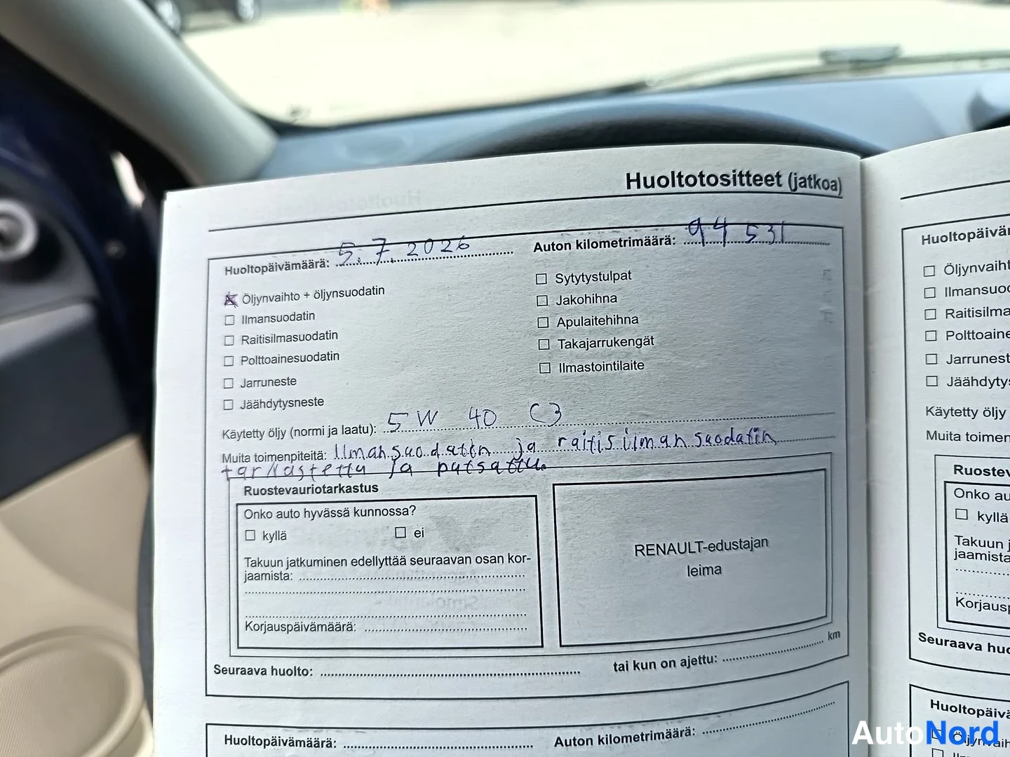 Muu: Huoltokirjan sivu auton sisällä kojelaudan edessä, huoltotosite päivämäärällä 5.7.2026, kilometrimäärä 94531, merkinnät ilmansuodattimen ja raitisilmasuodattime