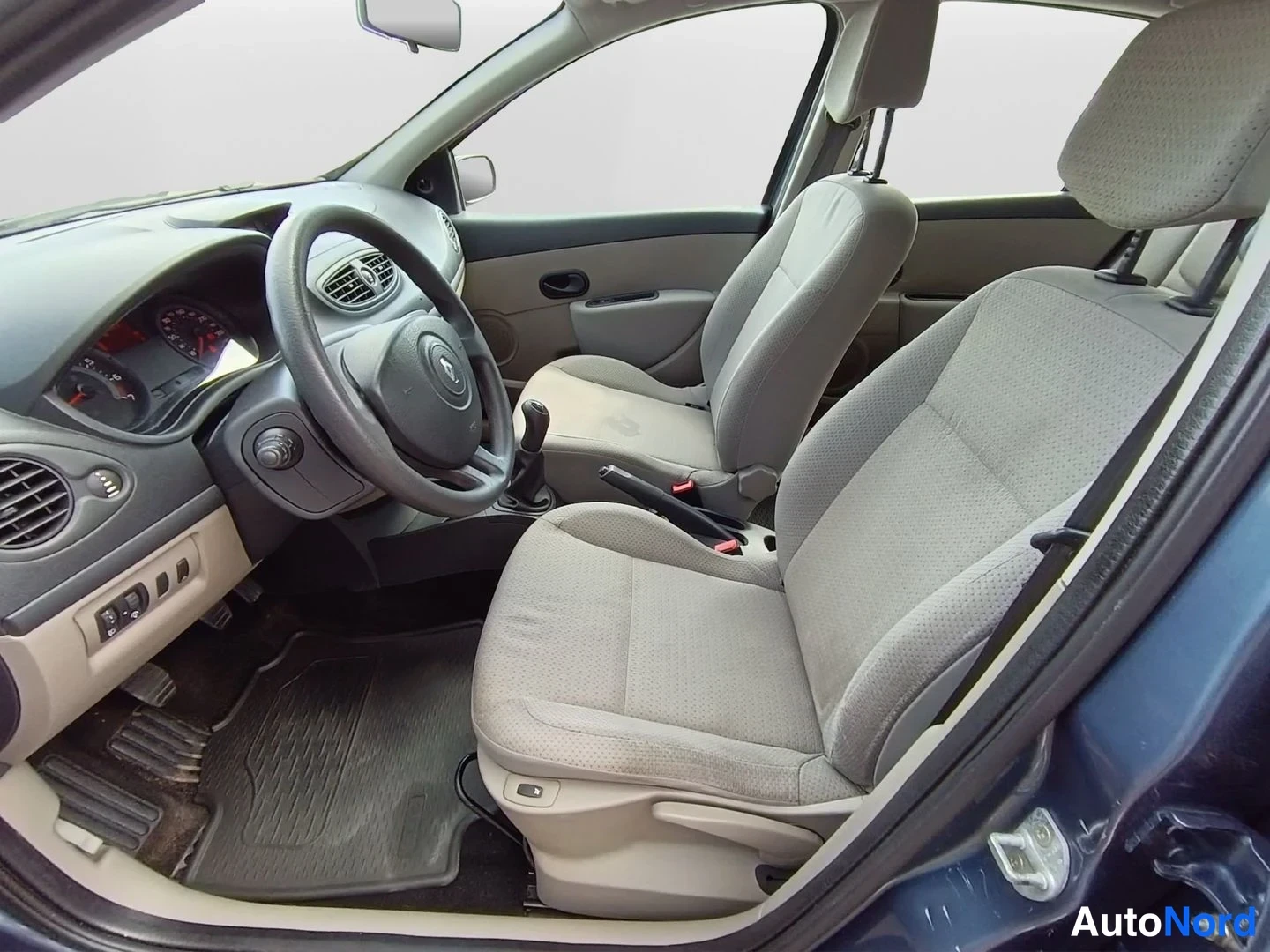 Sivulta: Renault Clio Confortin sisätilat: harmaat kangasistuimet, Renault-merkitty ohjauspyörä, kojelauta ja manuaalivaihteen vipu, kuljettajan ovi auki.