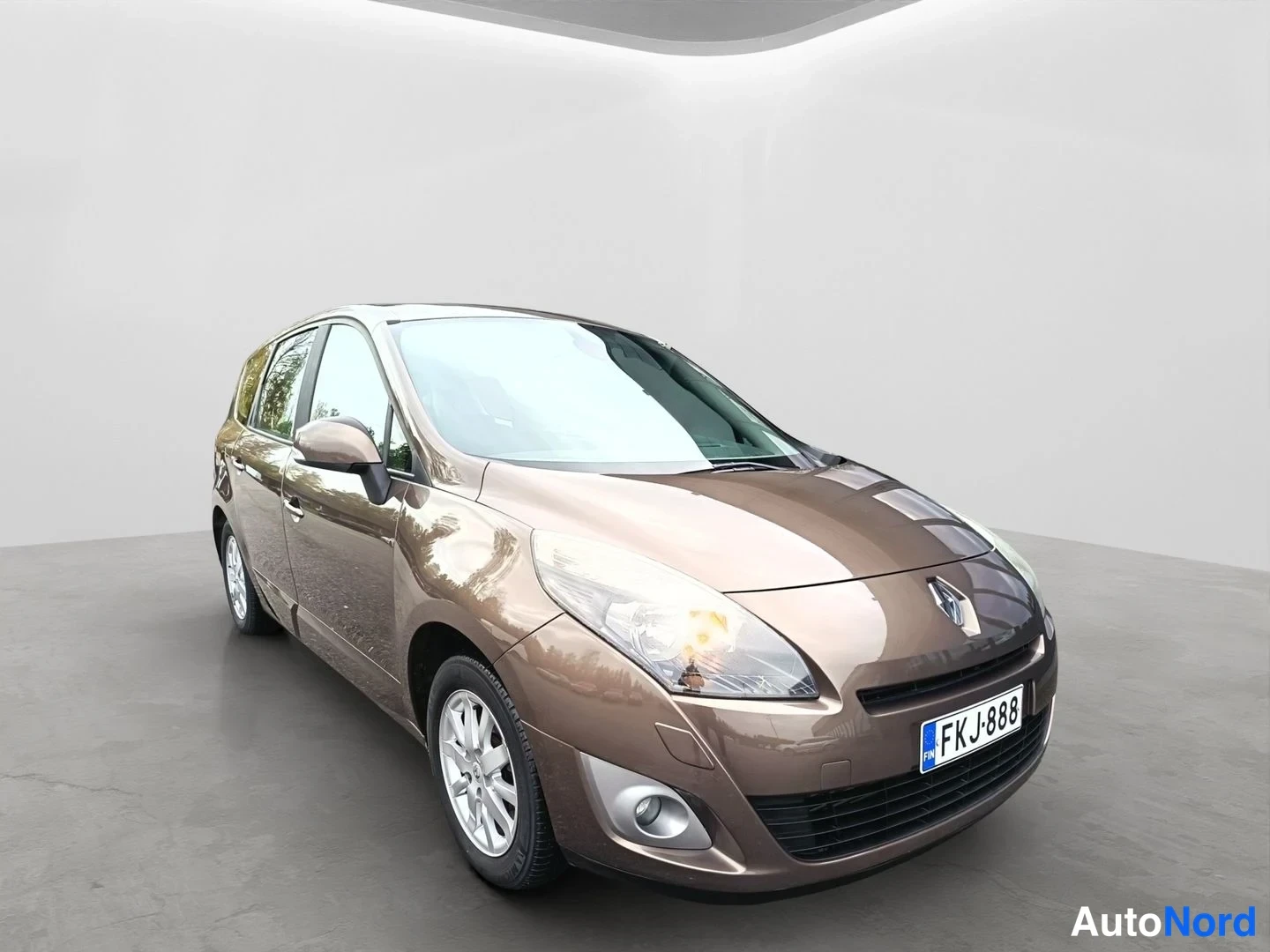 Etuviisto, nokka oikealle: Ruskea Renault Grand Scenic kuvattuna etuviistosta, keula osoittaa oikealle. Näkyvissä kylki, etuovet, sumuvalo ja alumiinivanteet; rekisterikilpi.