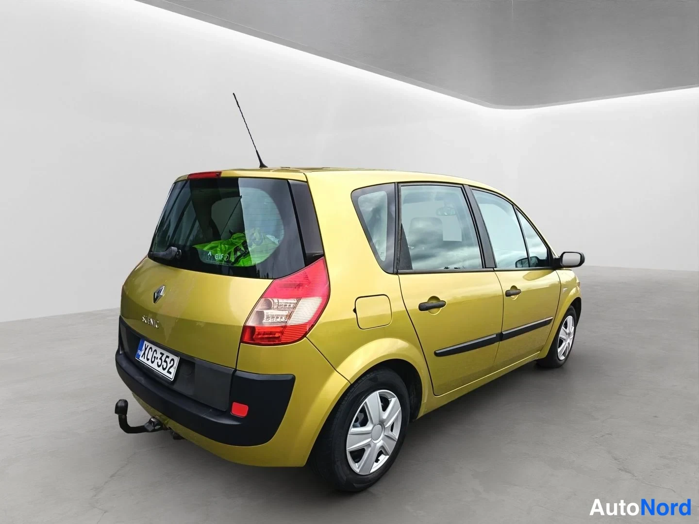 Takaviisto: Keltainen Renault Scenic kuvattuna takaa viistosti; takavalot, takaluukki, rekisterikilpi, kattiantenni ja vetokoukku näkyvät.