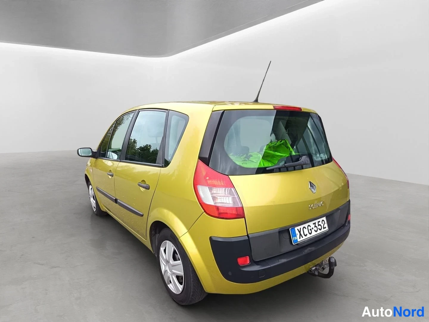 Takaviisto: Keltainen Renault Scenic kuvattuna takaosan ja vasemman kyljen nurkasta studiossa; näkyvissä takavalot, Renault-merkki, SCENIC-teksti, rekisterikilpi ja perävan