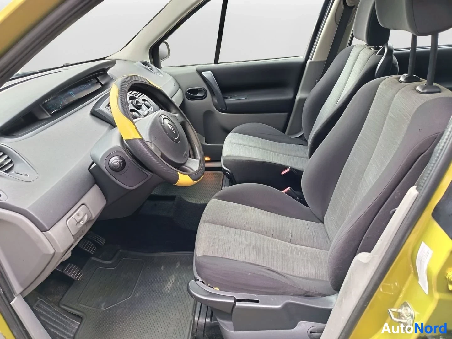 Muu: Renaultin sisätilat kuljettajan ovesta katsottuna: harmaa kojelauta, Renault-ohjauspyörä kelta-mustalla päällysteellä, kaksi tummaa kangasistuinta ja mattopesi