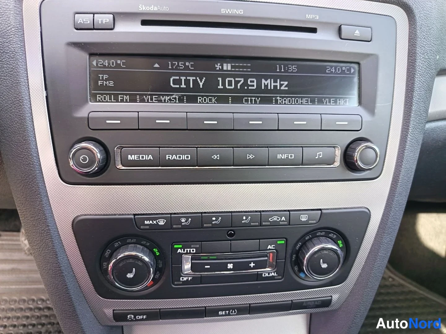 Muu: Kojelaudan keskiosan lähikuva: Škoda Swing -radio, näytöllä teksti CITY 107.9 MHz, ja alle kaksivyöhyke-automaattinen ilmastointi AC- ja AUTO-merkinnoilla.