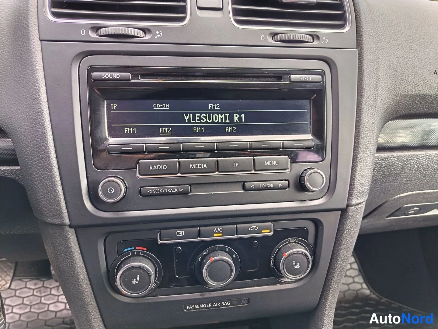 Etuistuimet: Keskikonsolin kuva. Autossa on radio-kasettisoitin, jonka LCD-näytössä lukee 'YLESUOMI R1'. Radioon kuuluvat fyysiset painikkeet radiolle ('RADIO'), mediapainik