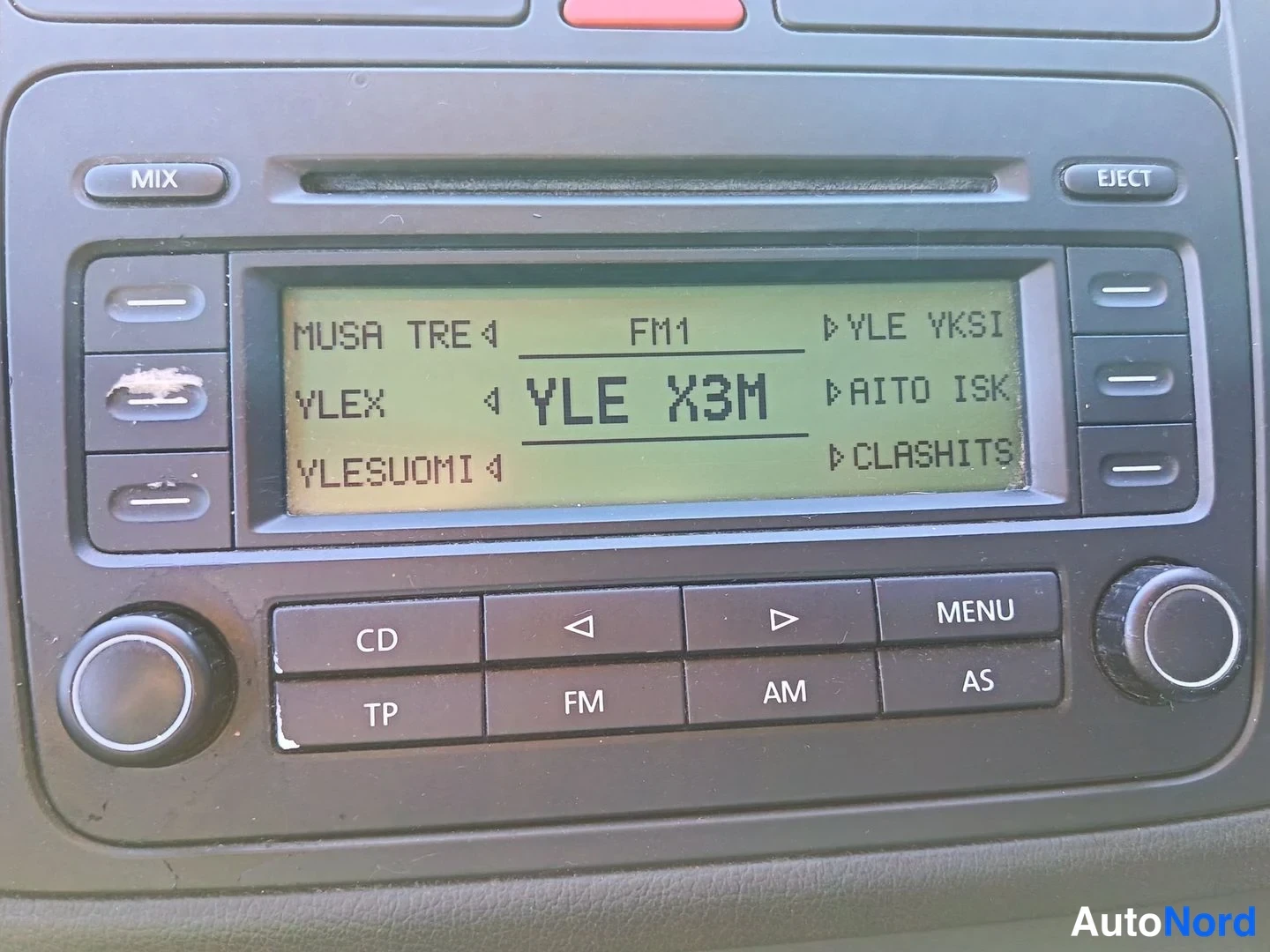 Yksityiskohta: Close-up of car stereo unit with LCD screen displaying YLE X3M radio station and menu items including YLE YKSI, RAITO ISK and CLASHITS. Buttons around display i