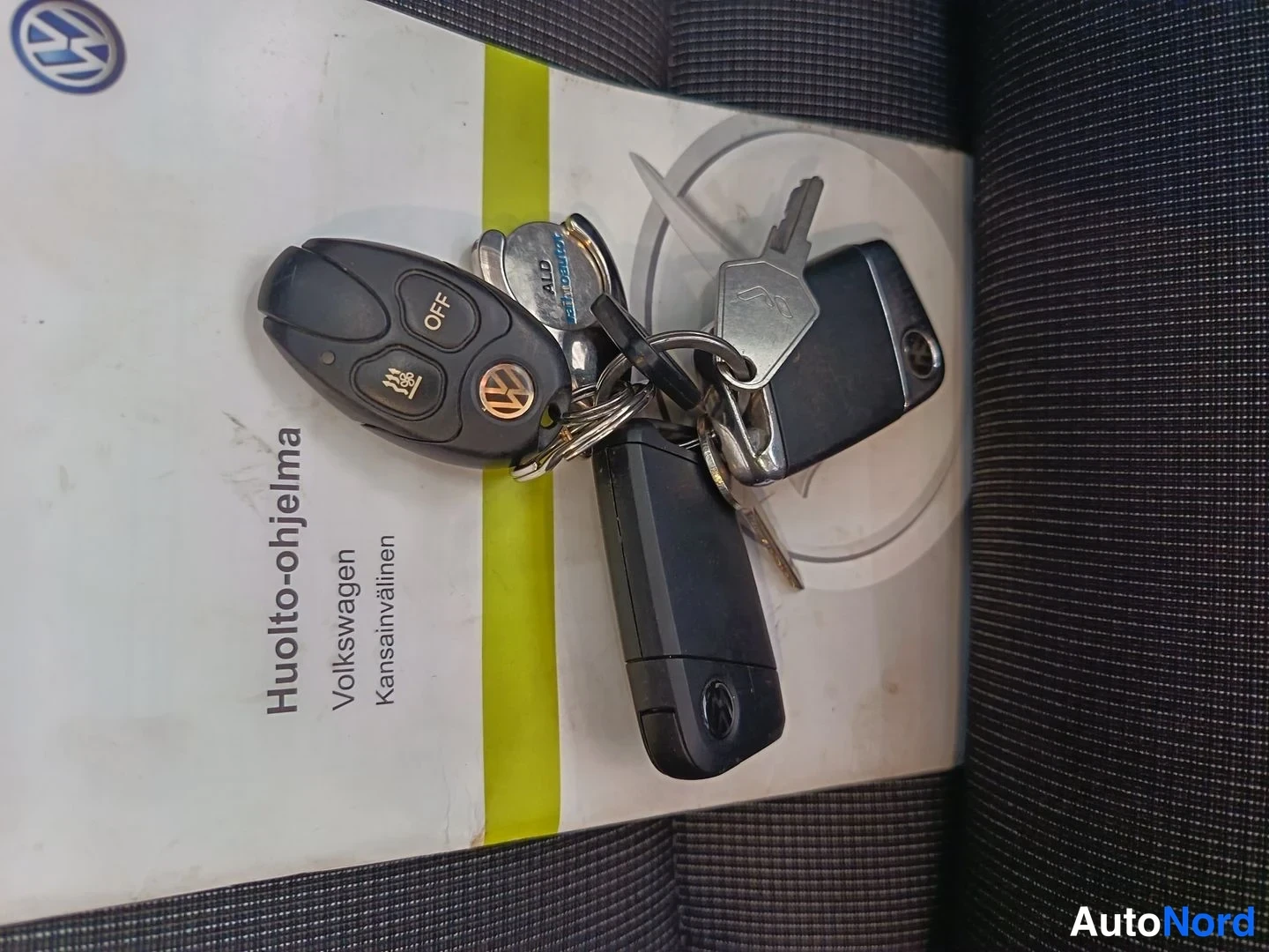 Yksityiskohta: A set of keys resting on a manual cover featuring the Volkswagen logo. The key fob is black with buttons labeled 'OFF' and an icon representing a heated seat.