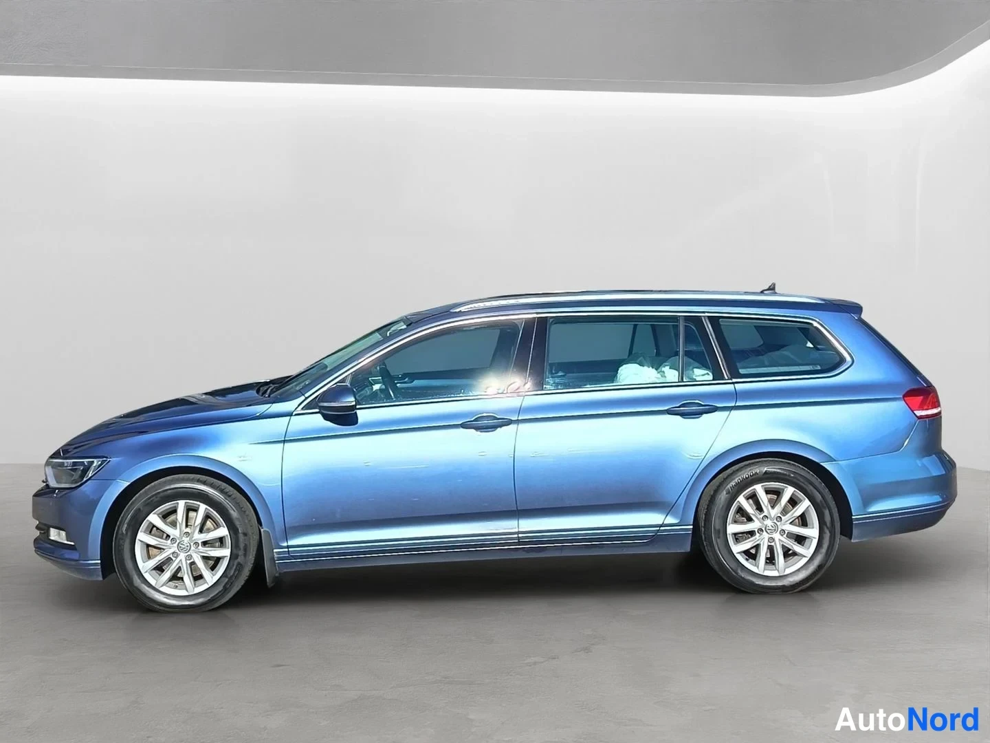 Sivulta: Side profile of a blue Volkswagen Passat Variant station wagon with silver alloy wheels, door handles, side steps and roof rails against a plain wall