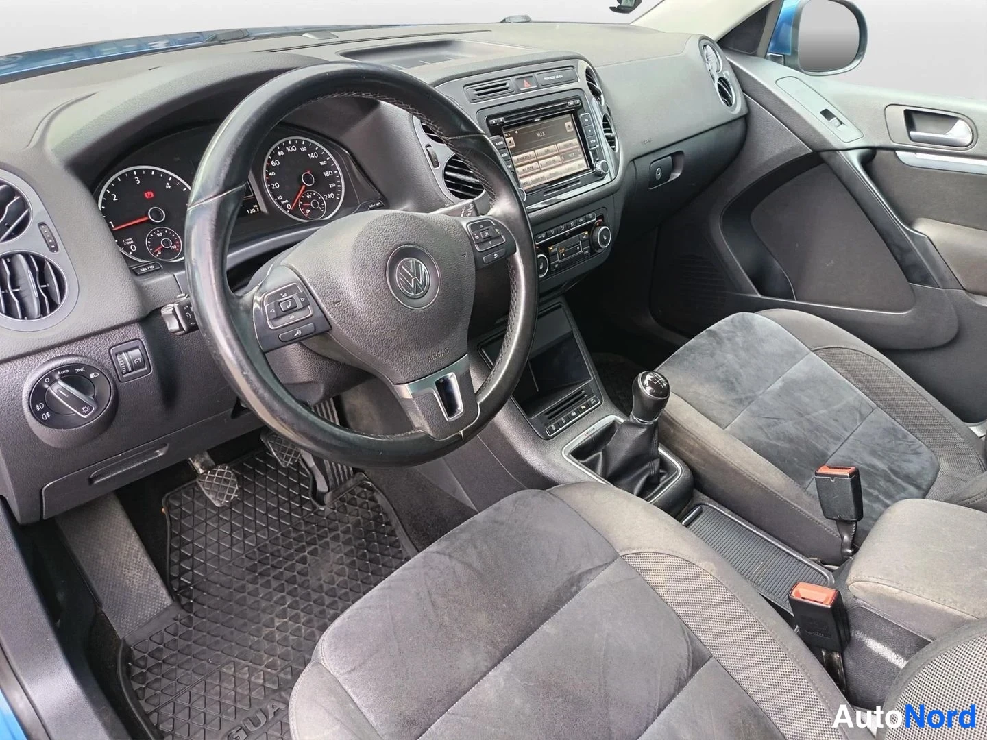 Edestä: Volkswagen Tiguanin kojelauta ja etuistuimet kuvattuna kuljettajan puolelta; näkyvät monitoimiohjauspyörä, mittaristo, radio, manuaalivaihto ja kangasistuimet.