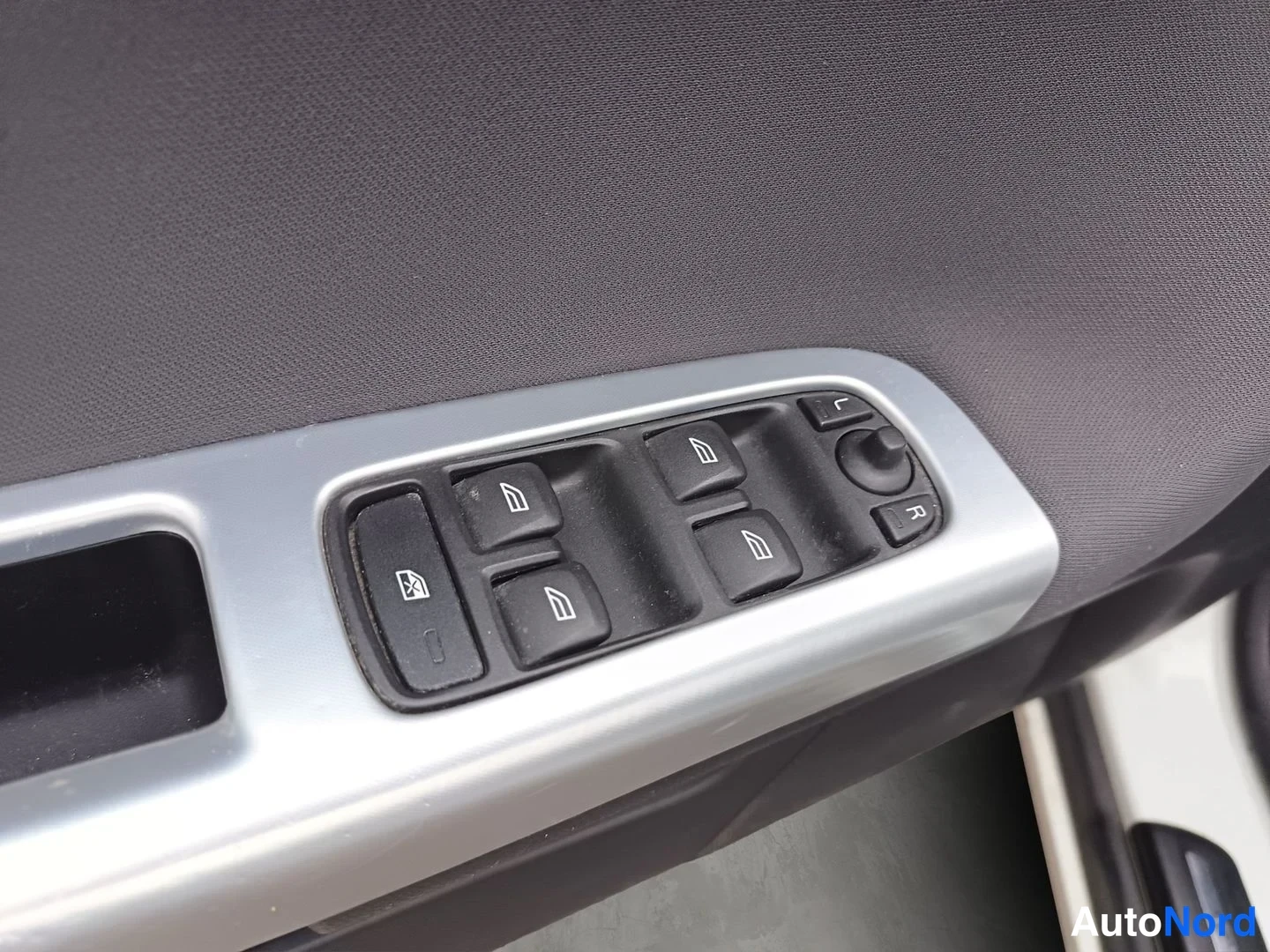 Yksityiskohta: Close-up of the driver-side front door control panel featuring window switches with icon labels (auto-lock), power mirror adjustment knob with directional marki