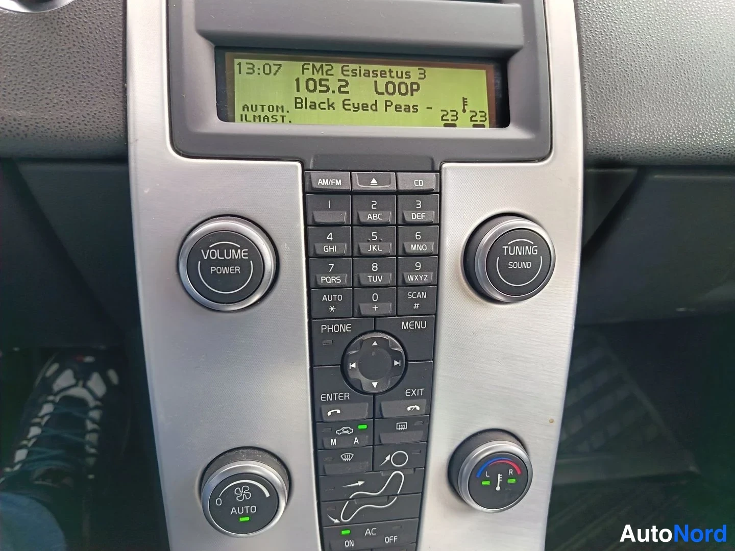 Yksityiskohta: Keskikonsolin radio- ja ilmastointipaneeli lähikuvassa: näytöllä lukee '105.2 LOOP', säätimet on merkitty VOLUME ja TUNING, ja paneelissa lukee 'AUTOM. ILMAST.'