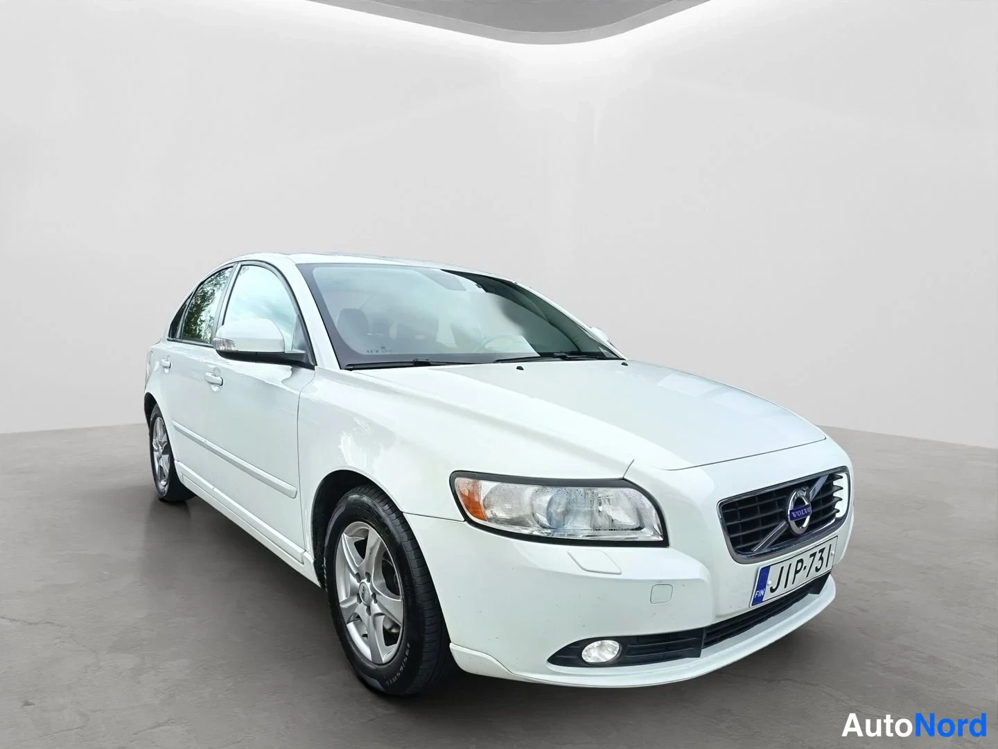 Etuviisto, nokka oikealle: Front three-quarter shot of the other side of a white Volvo sedan. Visible equipment: alloy wheels.