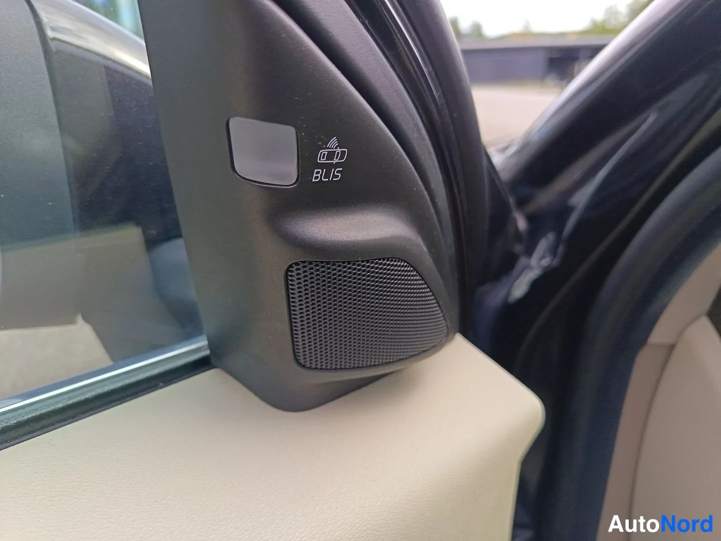 Yksityiskohta: Close-up of black side-mirror housing showing a BLIS blind-spot indicator button above a speaker grille.