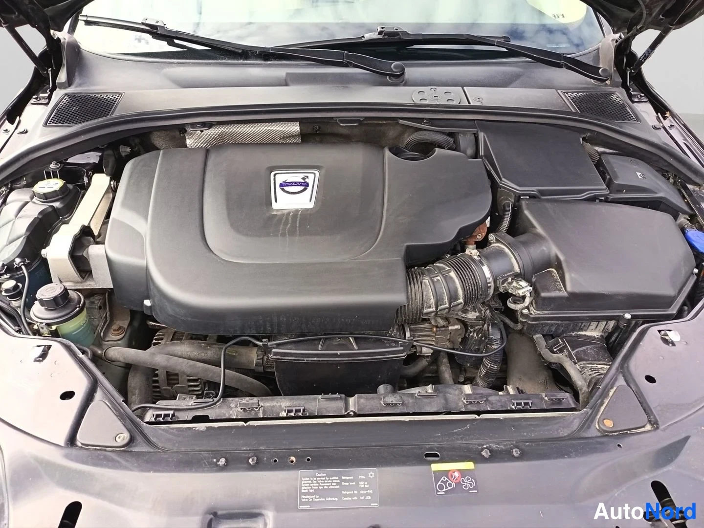 Moottoritila: Engine bay view under open hood showing black plastic engine cover with Volvo badge at center, hoses and fluid reservoirs around it.
