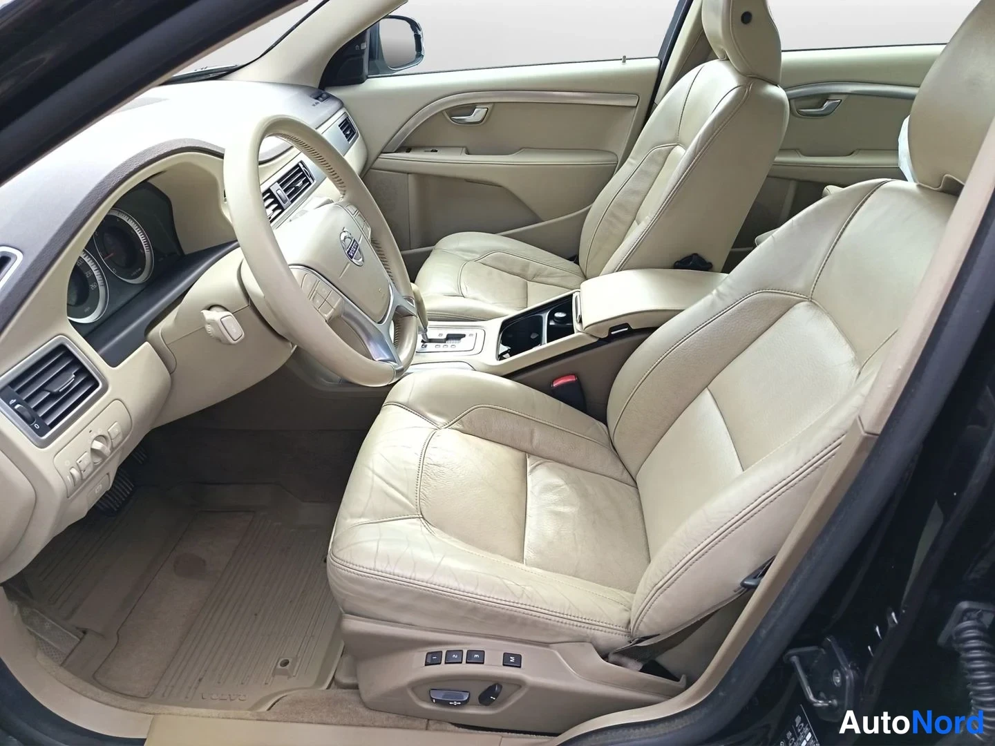 Ohjaamo: Interior view from the open front passenger door showing the driver's seat and front passenger seat. The seats are upholstered in beige leather with multi-way p