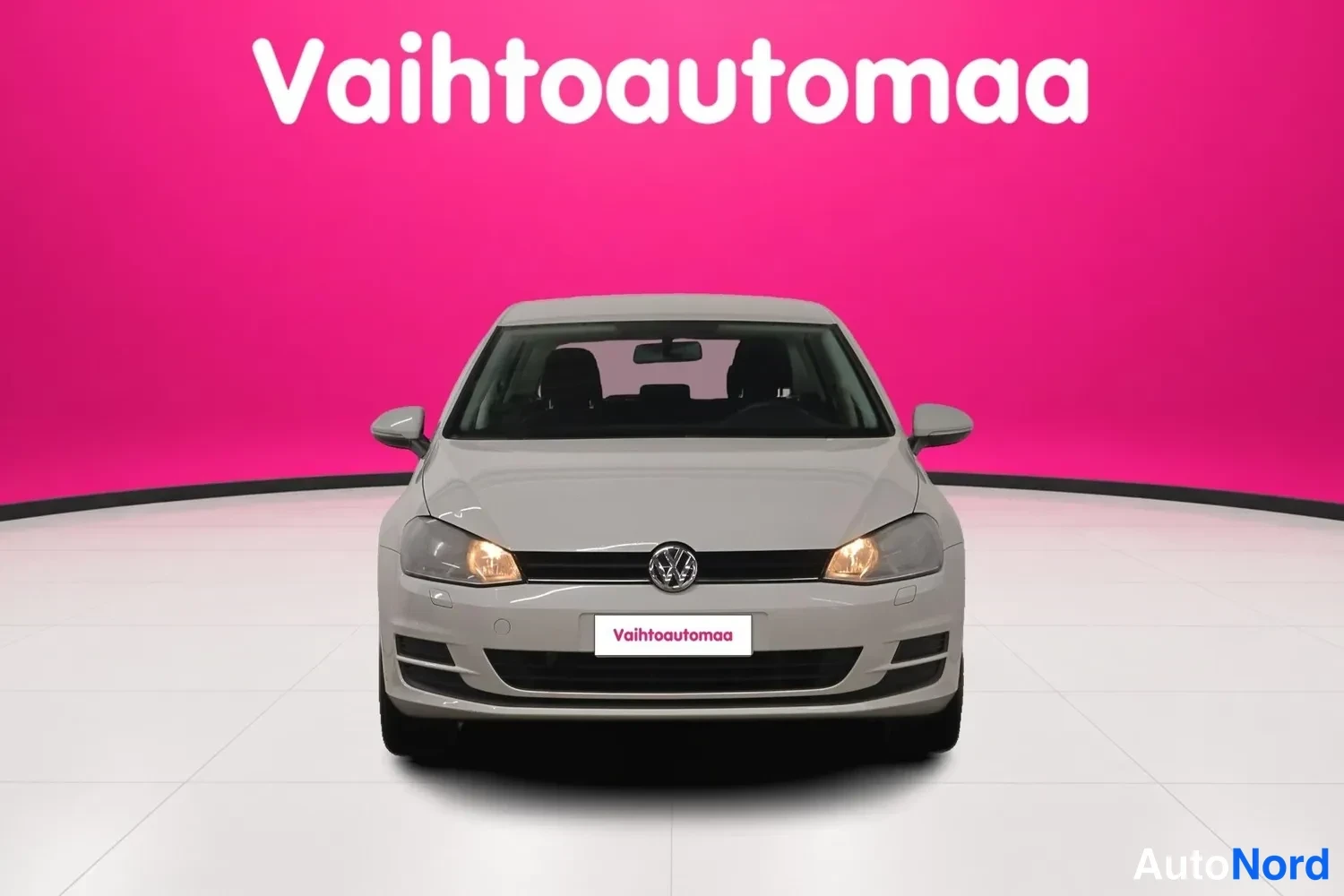 Edestä: Suora etukuva valkoisesta hatchbackista, johon on merkitty Vaihtoautomaa-teksti sekä kyltissä että taustalla olevassa ruudussa; etupuskurissa näkyy etusumunvalo